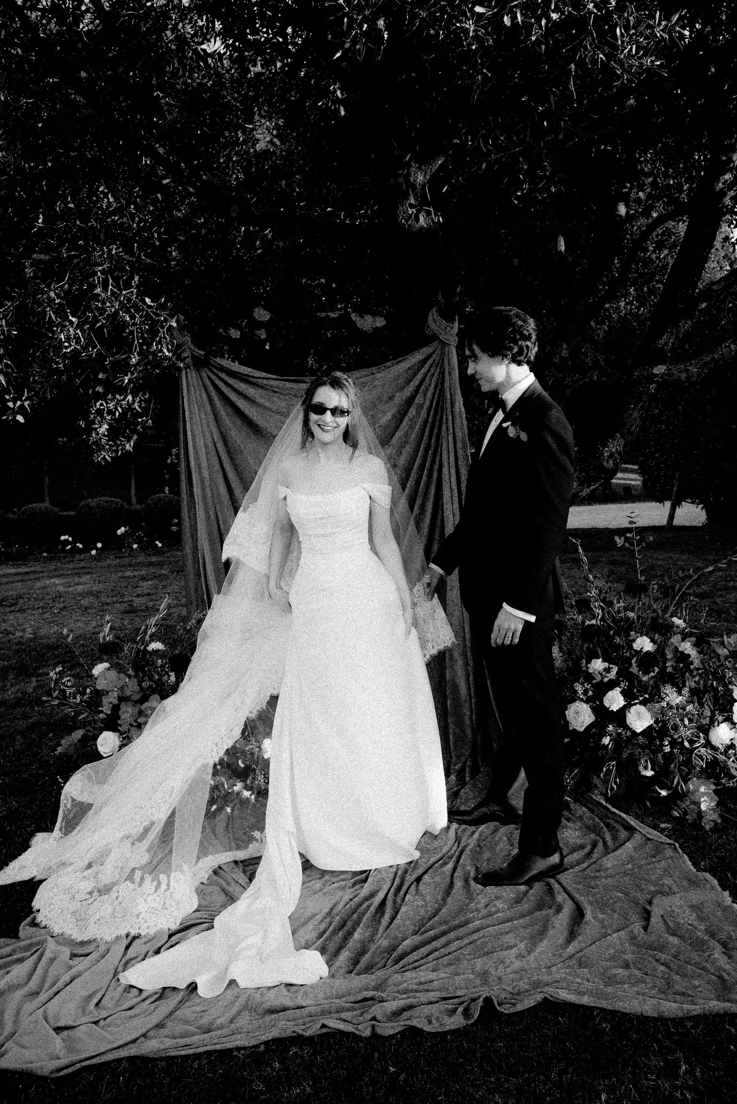 Black and white photo of a bride in a white gown and veil with sunglasses standing with a groom in a tuxedo outdoors.