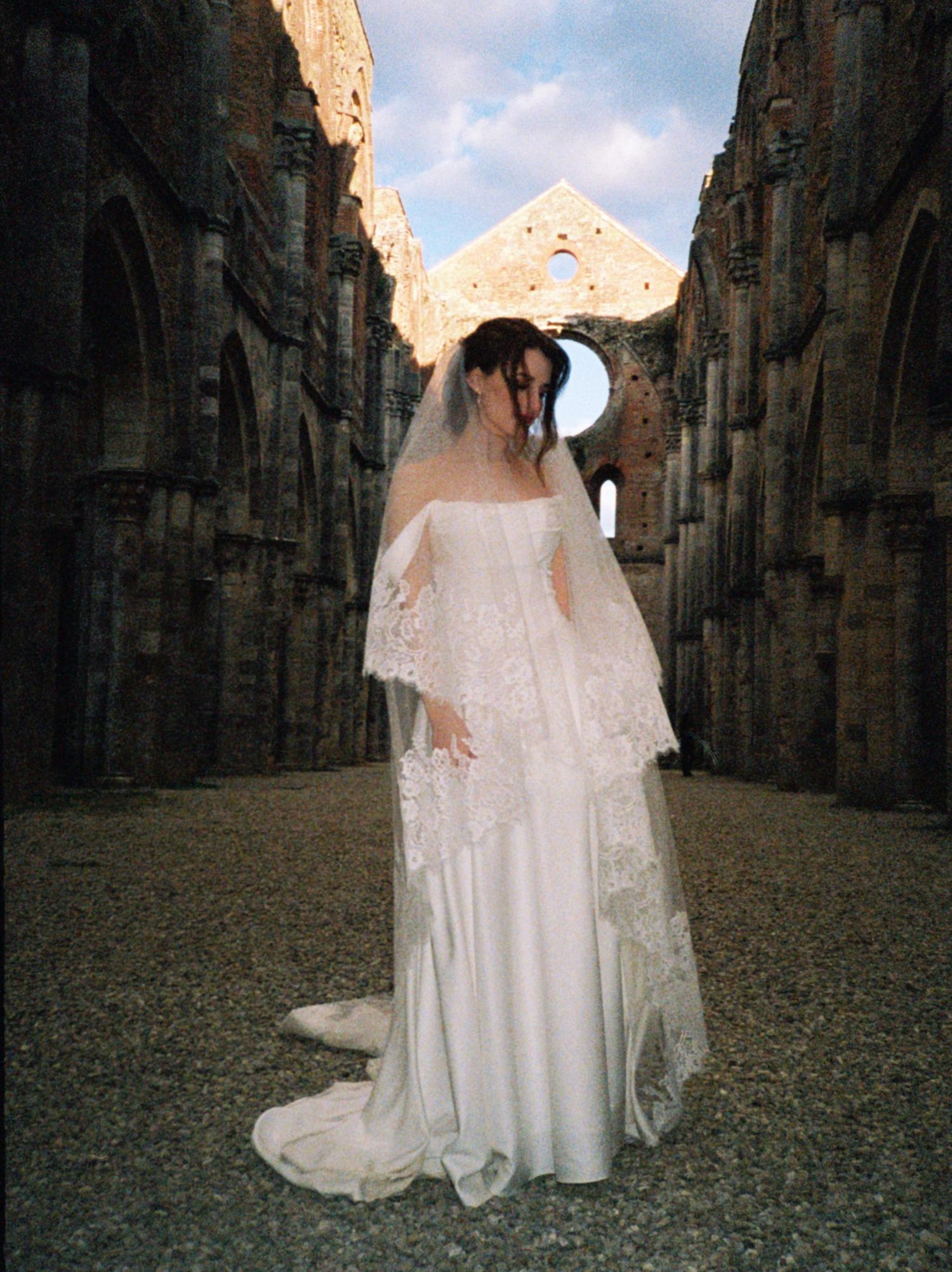 A figure in a flowing white wedding dress and veil stands in the stone ruins of an ancient abbey.