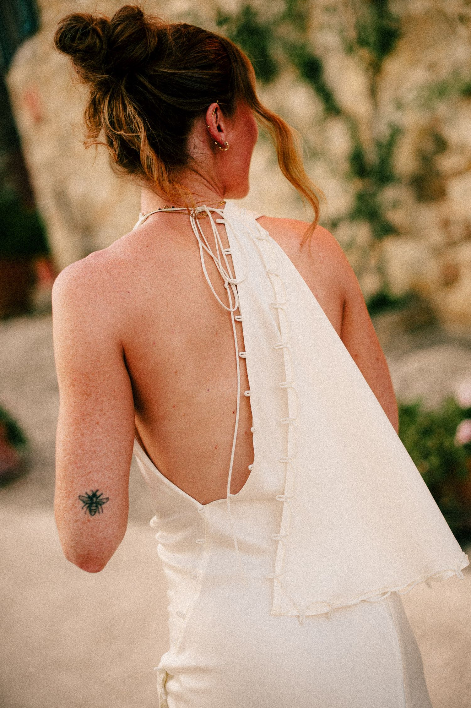 Woman in backless white halter dress with bee tattoo on arm, showing elegant lace-up back detail.
