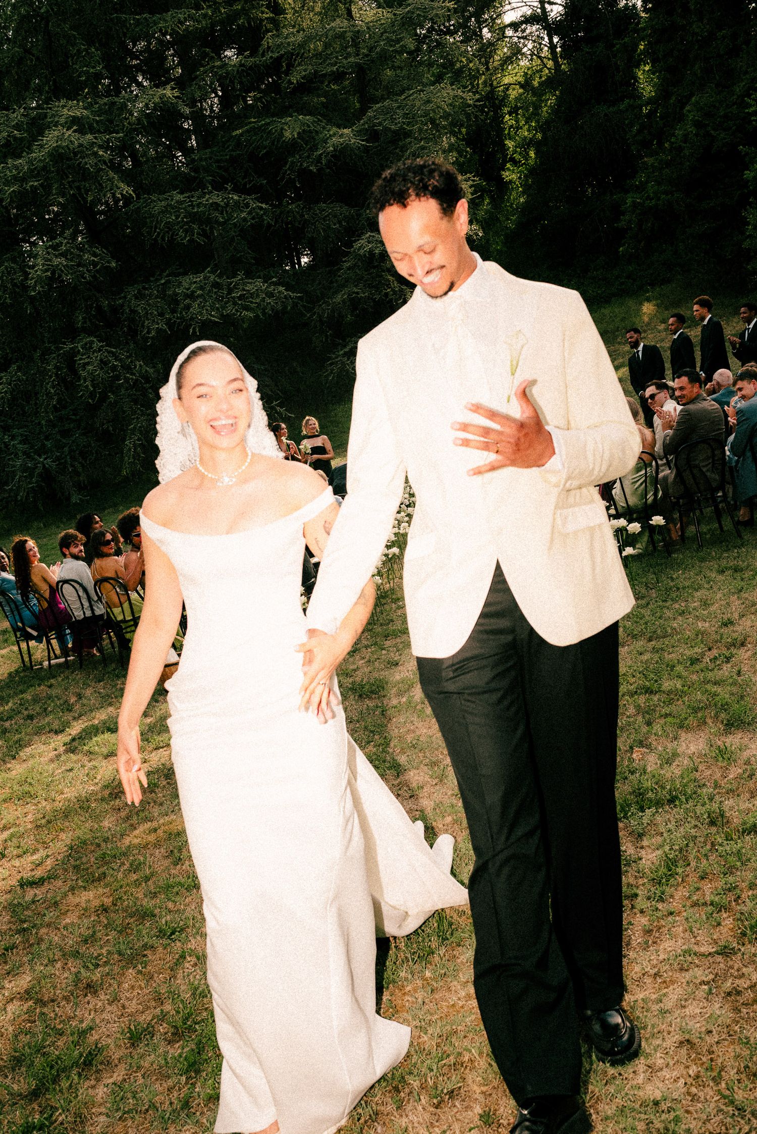 A smiling bride in white gown and groom in white jacket walk together at an outdoor wedding ceremony.