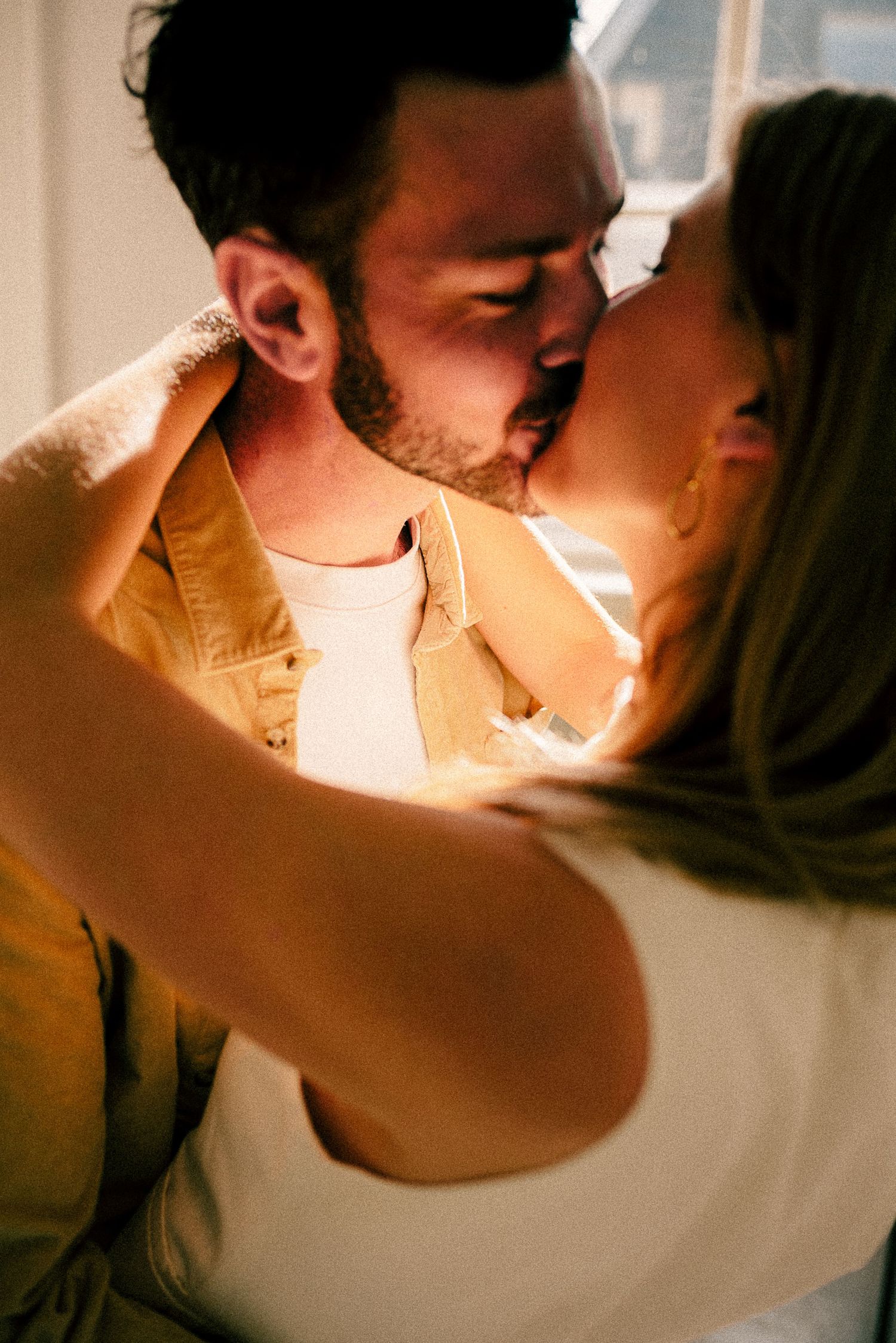 A couple sharing a romantic kiss, the man in a yellow jacket embracing a woman in warm golden light.