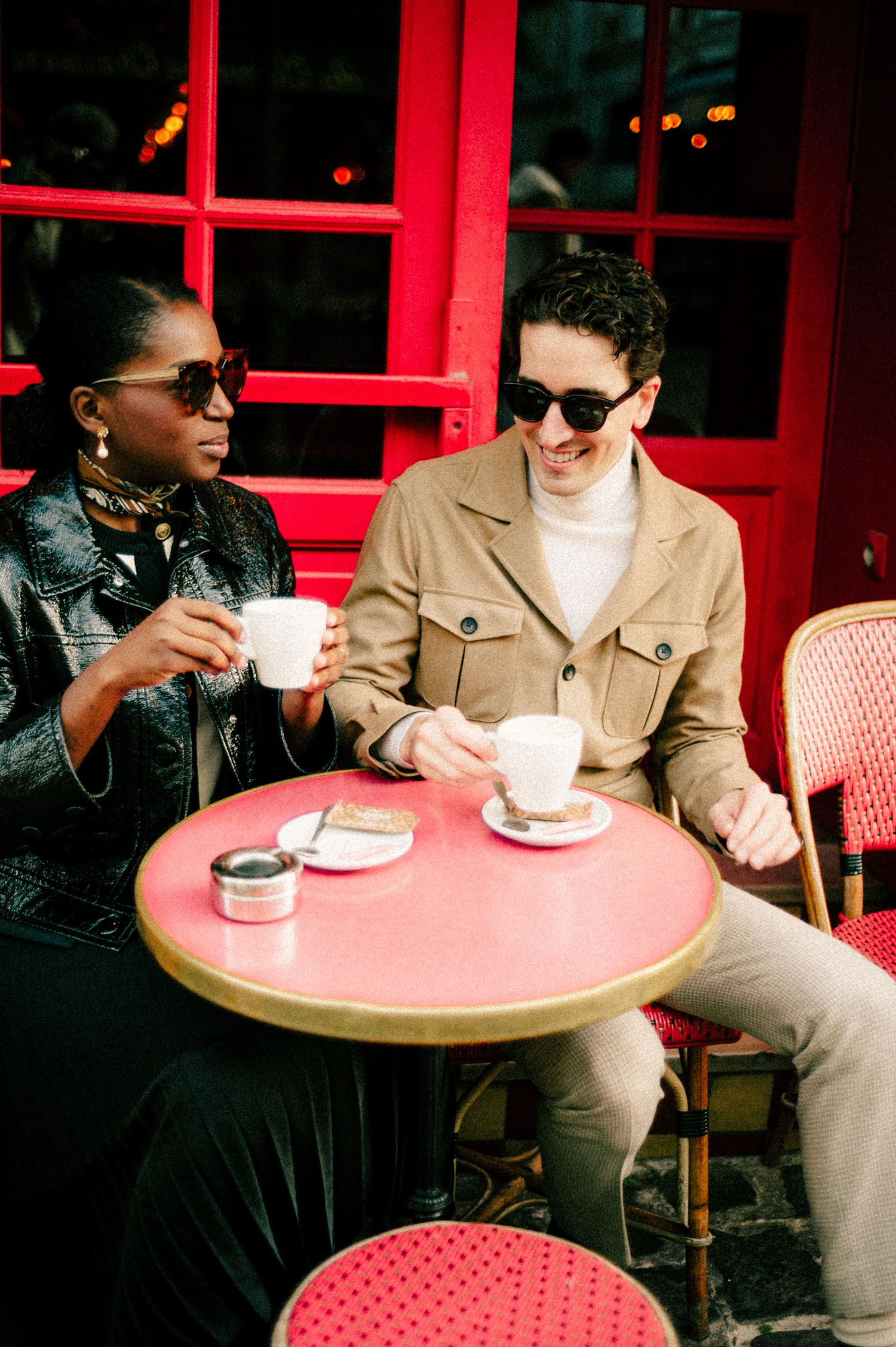 Two friends enjoy coffee at a red Parisian cafe table outdoors, wearing sunglasses and stylish outfits.