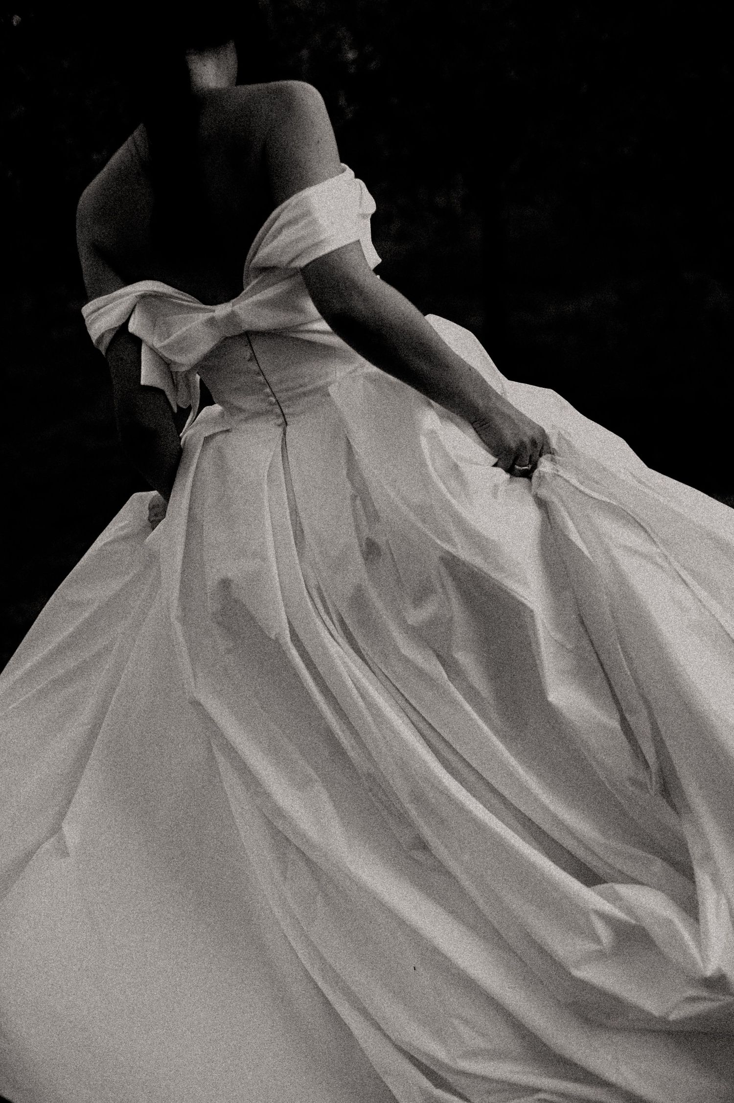 Black and white photo of a woman in an elegant off-shoulder ball gown, gracefully holding the flowing white skirt.