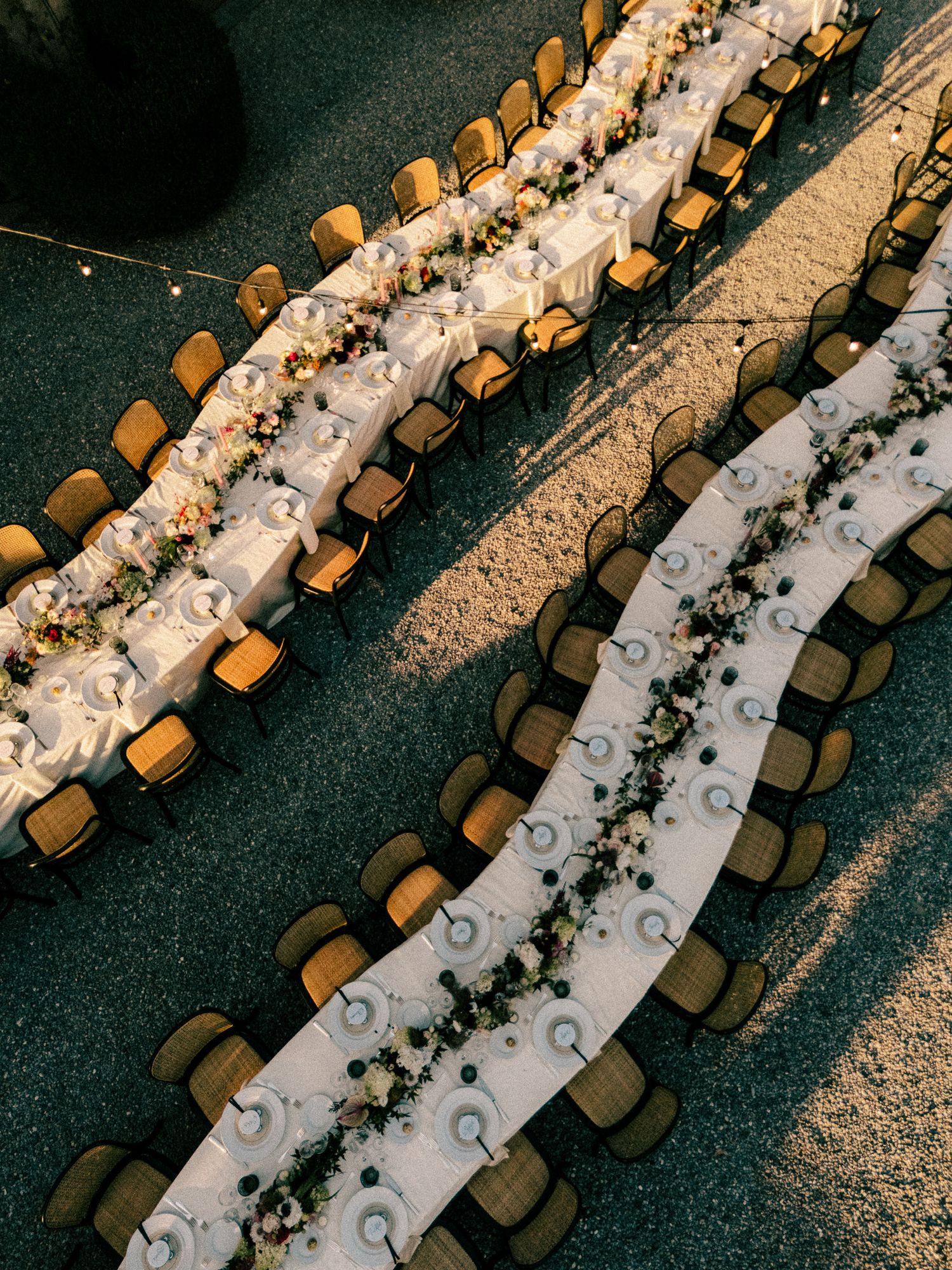 Aerial view of elegant outdoor wedding reception with long white tables, floral runners, and gold chairs.