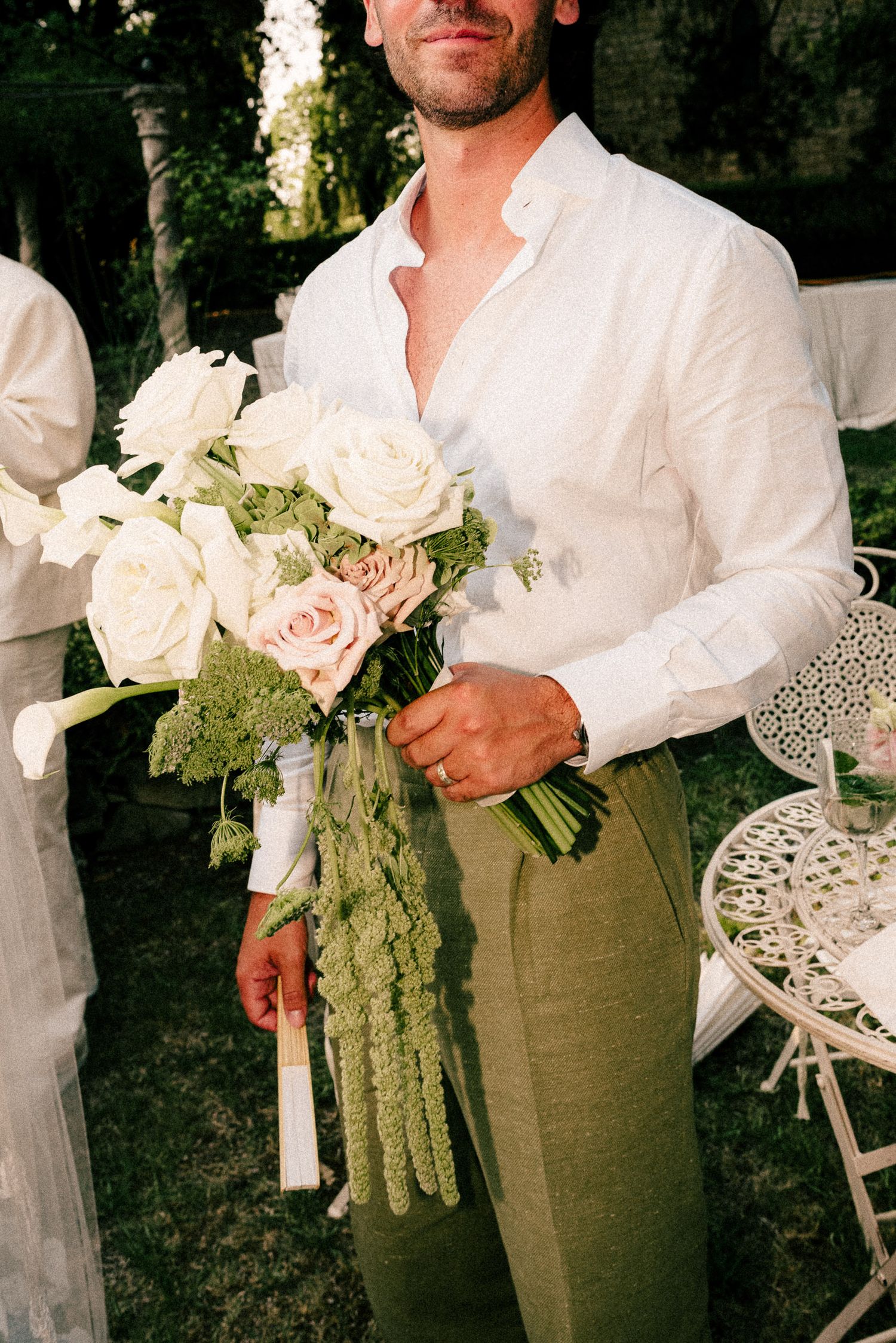 Man in white linen shirt and green trousers holding elegant bridal bouquet of white and pink roses outdoors.