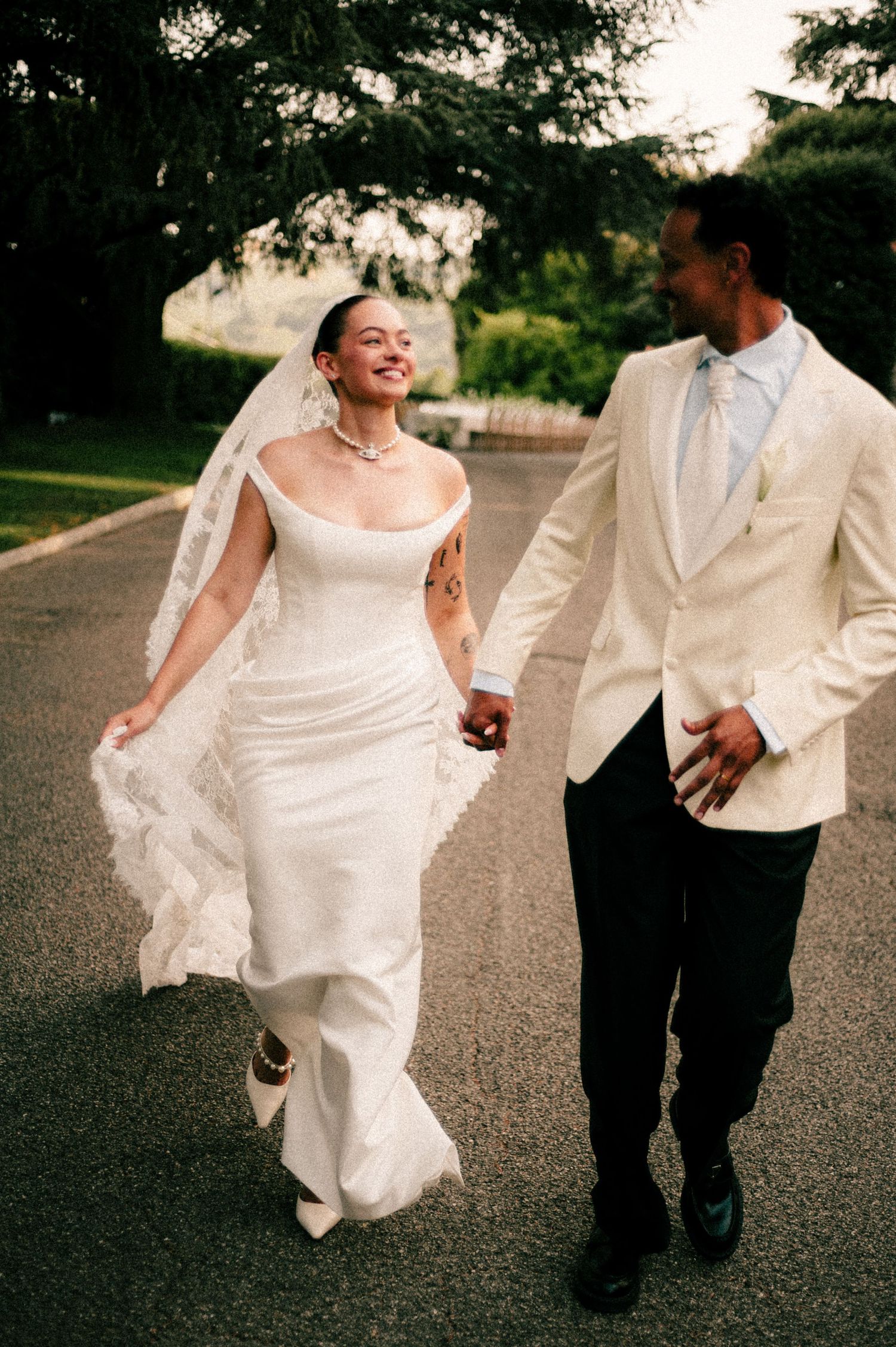 Bride and groom walking hand in hand outdoors, she in a white off-shoulder gown with veil, he in a cream tuxedo jacket.