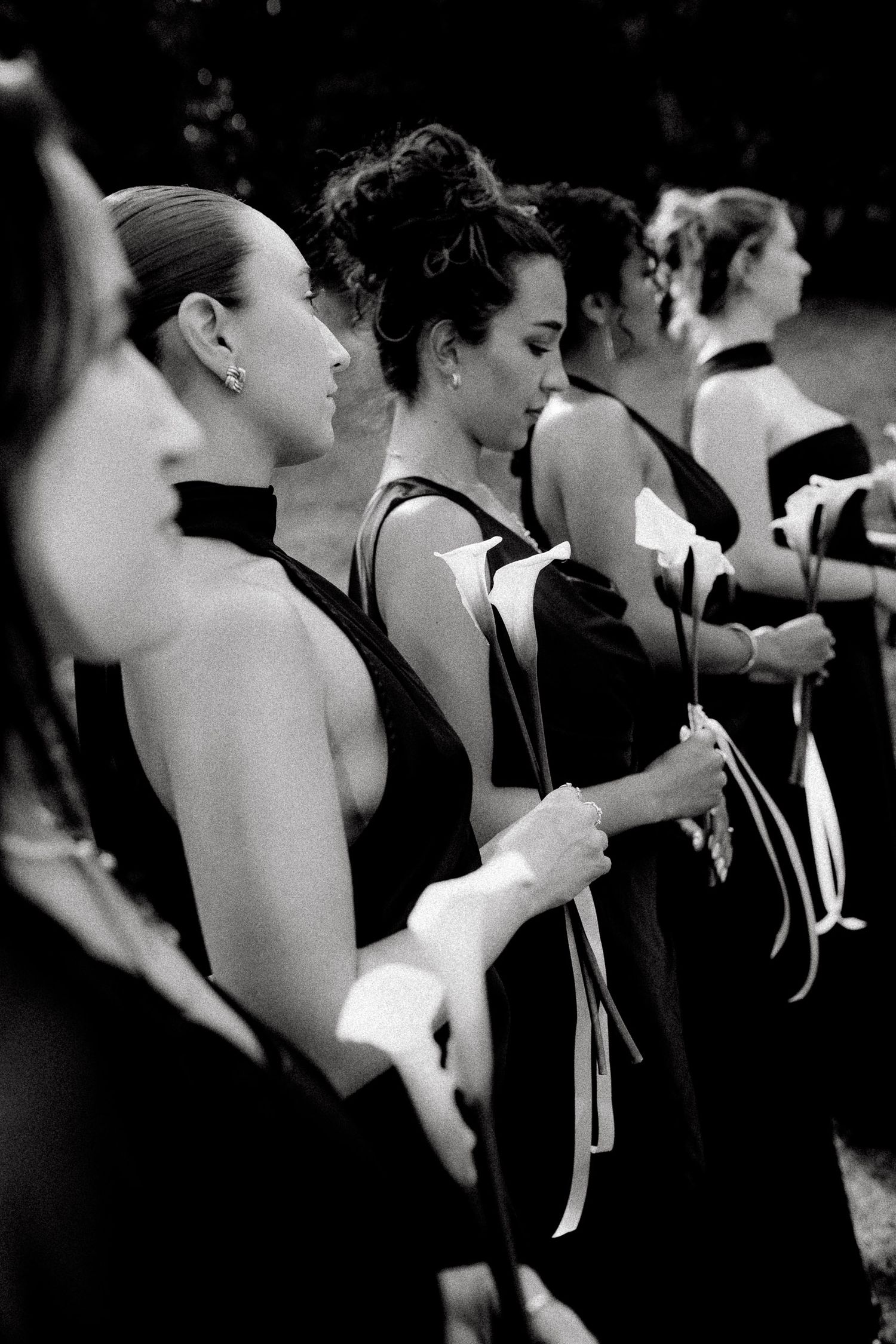 Bridesmaids in elegant black halter gowns holding white ribbon bouquets stand in a row at an outdoor wedding ceremony.