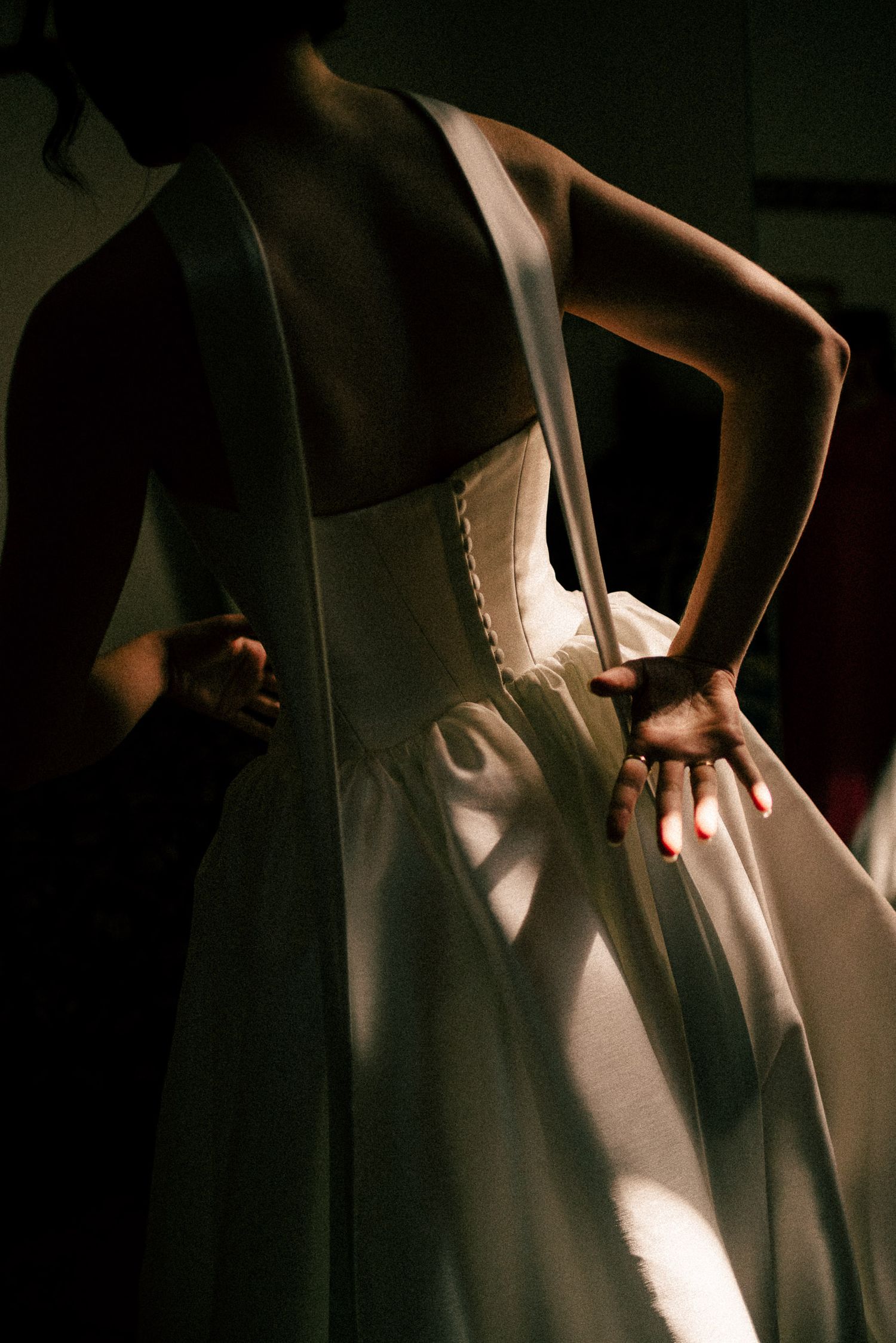 A bride in a white wedding gown being laced up from behind, with dramatic shadows and warm light.