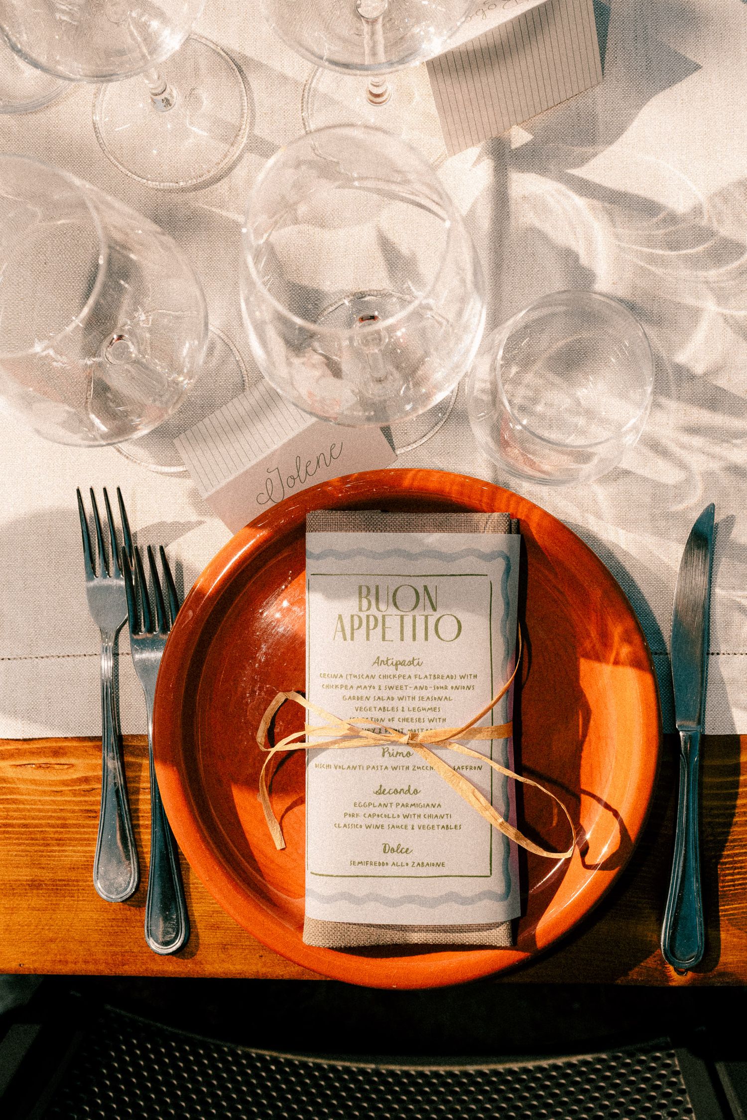 Elegant Italian place setting with orange plate, Buon Appetito menu, wine glasses, and silverware on linen tablecloth.