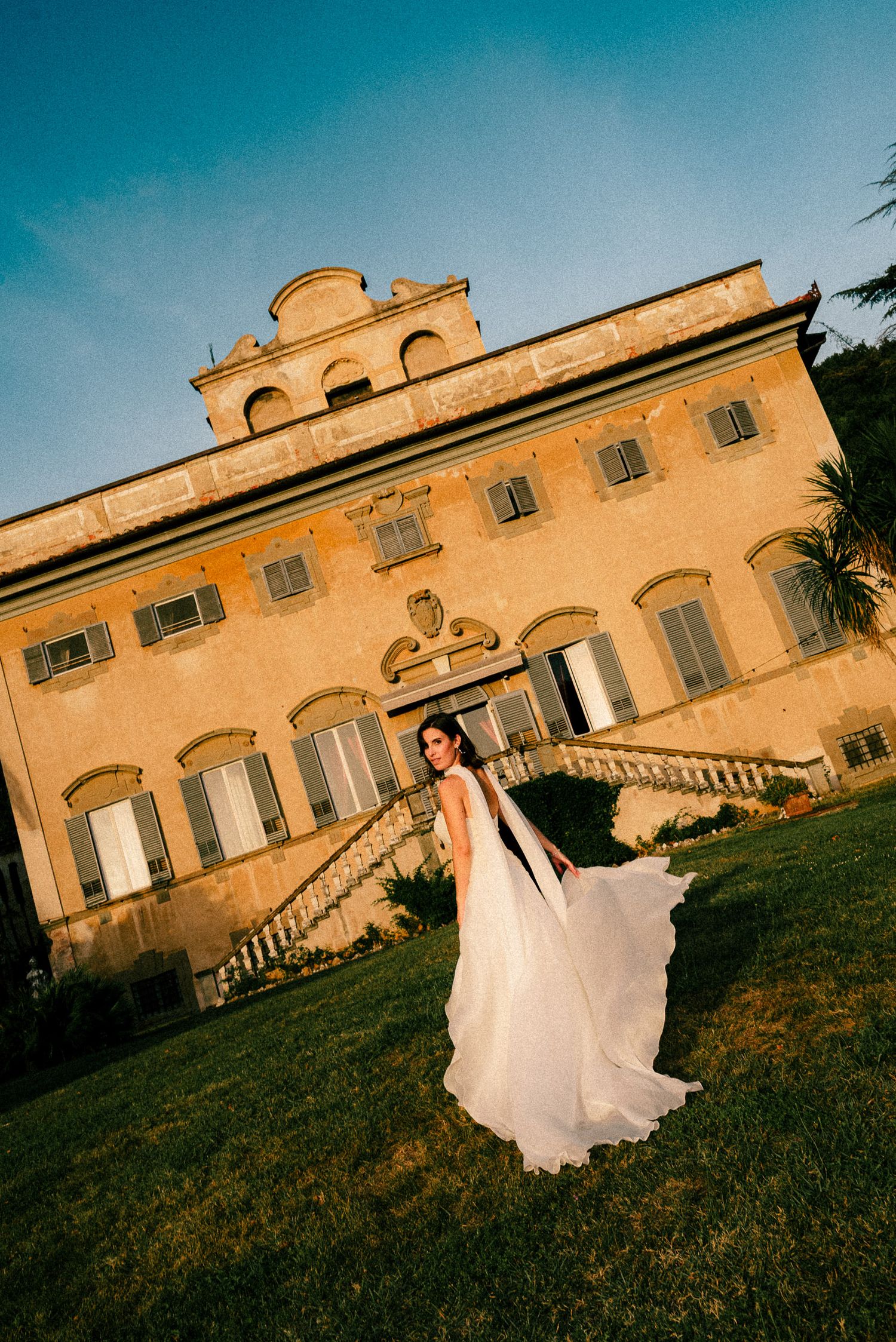 Bride in flowing white gown poses before elegant Italian villa on lush green lawn at golden hour.