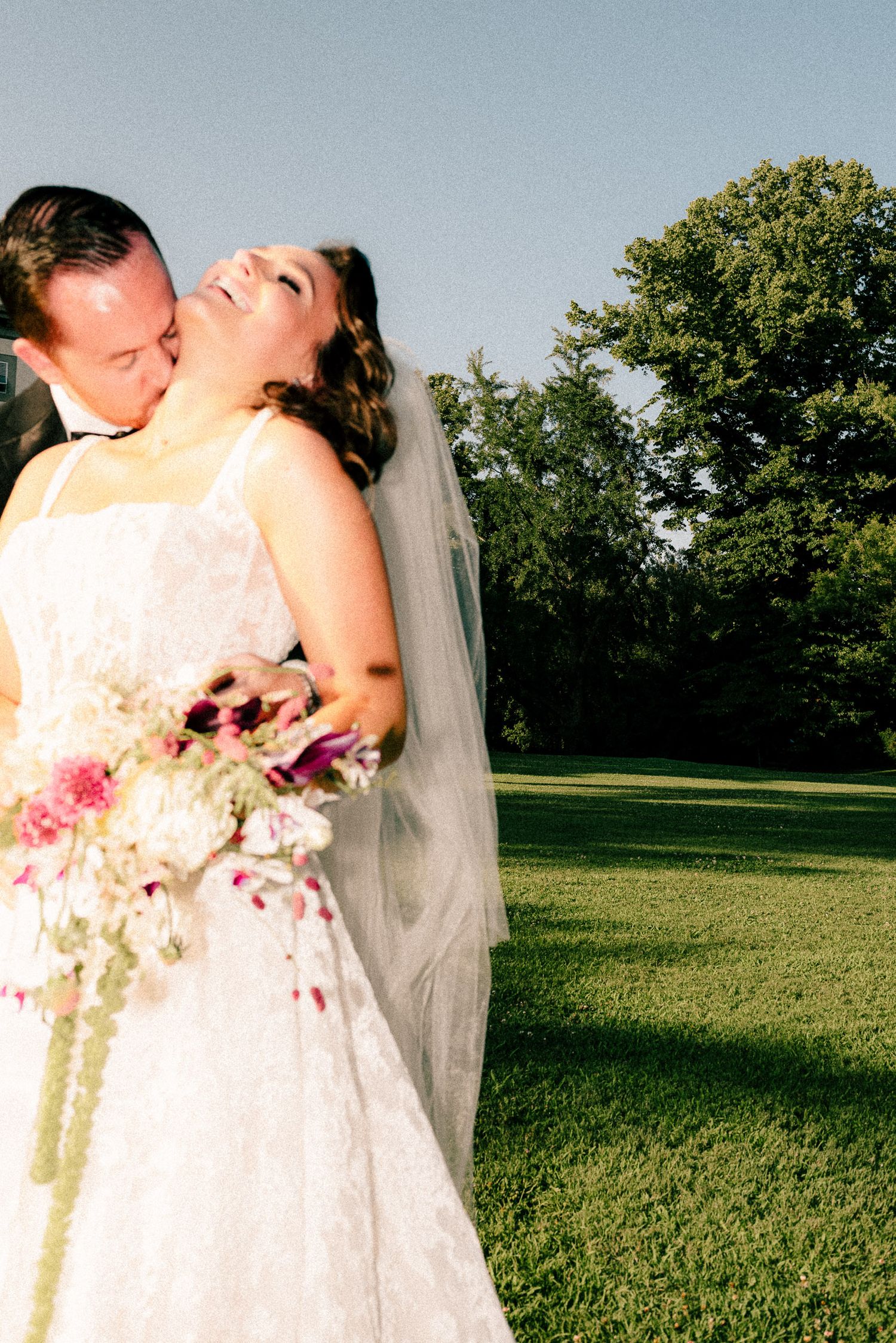 Groom kissing bride on cheek outdoors, bride laughing holding pink floral bouquet in white wedding dress with veil.