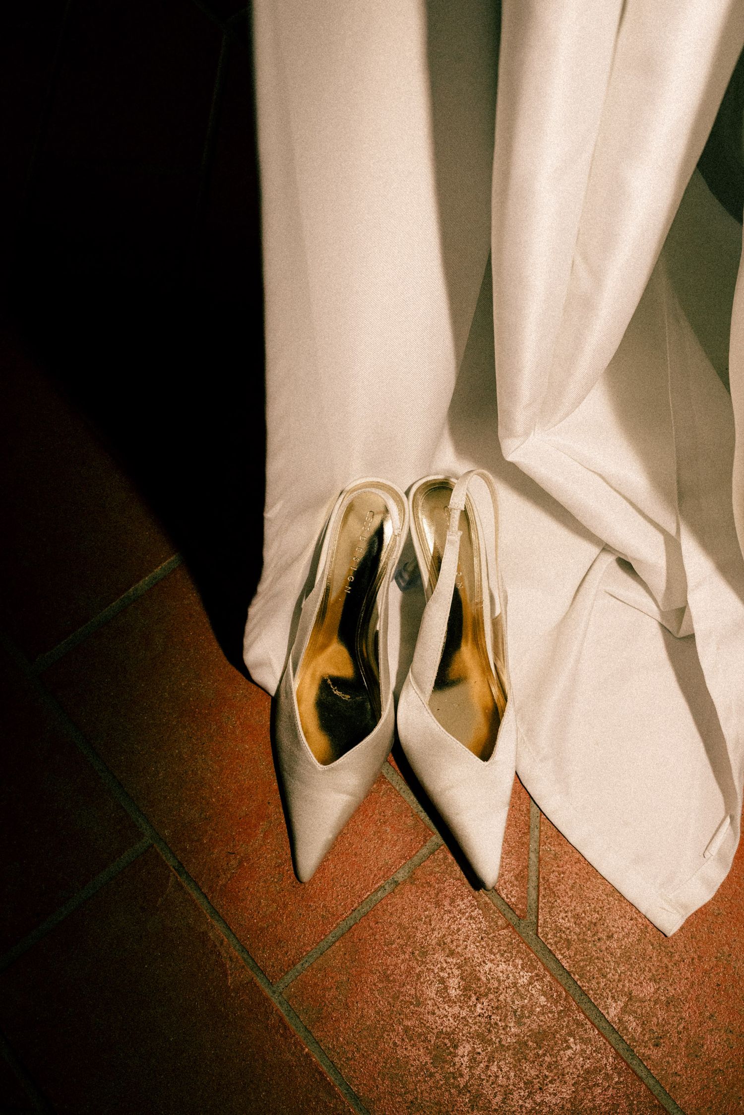 Elegant white bridal heels with gold insoles placed on terracotta tile floor beside a flowing white wedding dress.