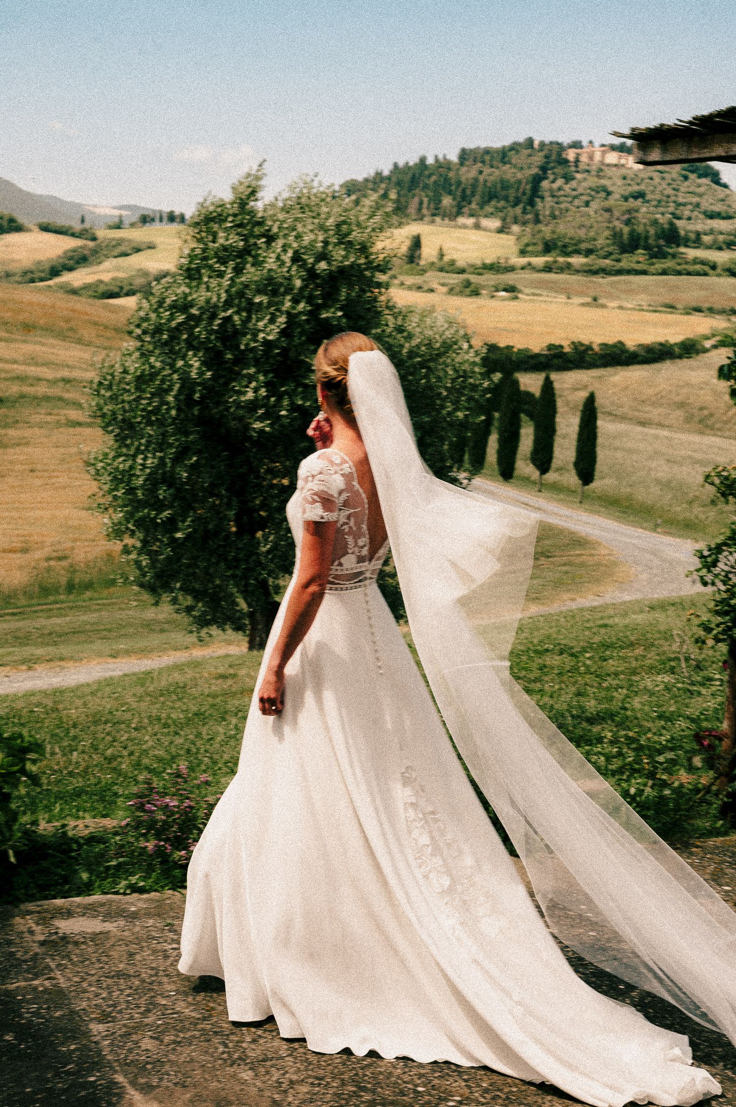 Bride in a flowing white gown with lace back and long veil stands overlooking Tuscan countryside hills.