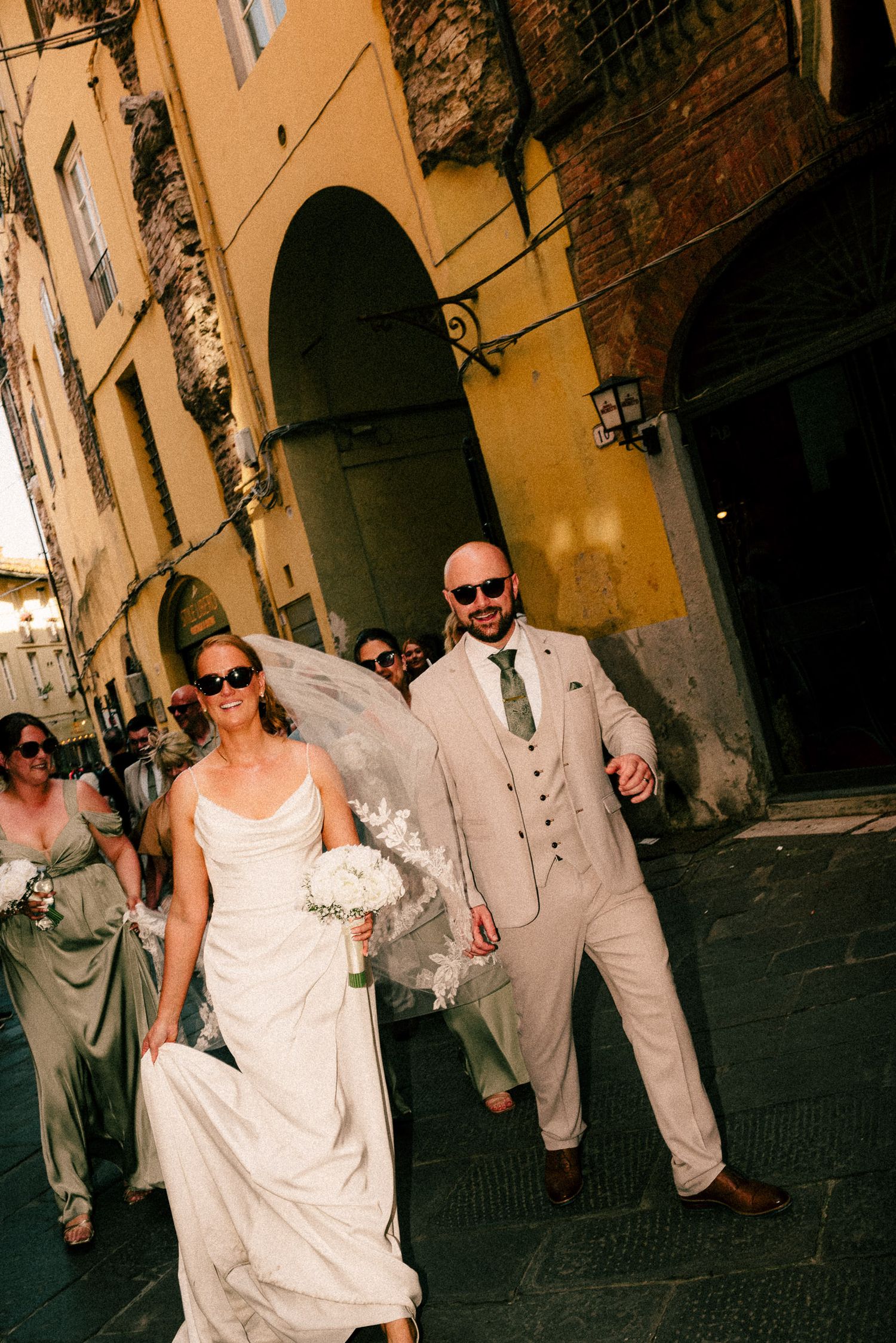 Bride and groom walking through historic Italian streets in wedding attire, with bridesmaids following behind.