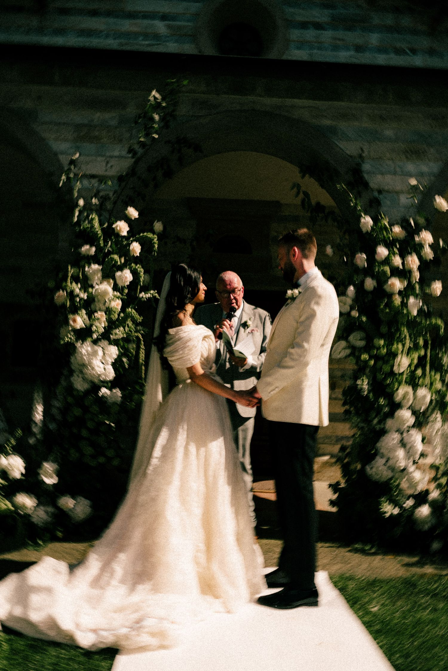Bride and groom exchange vows under a floral arch adorned with white roses at an elegant outdoor wedding ceremony.