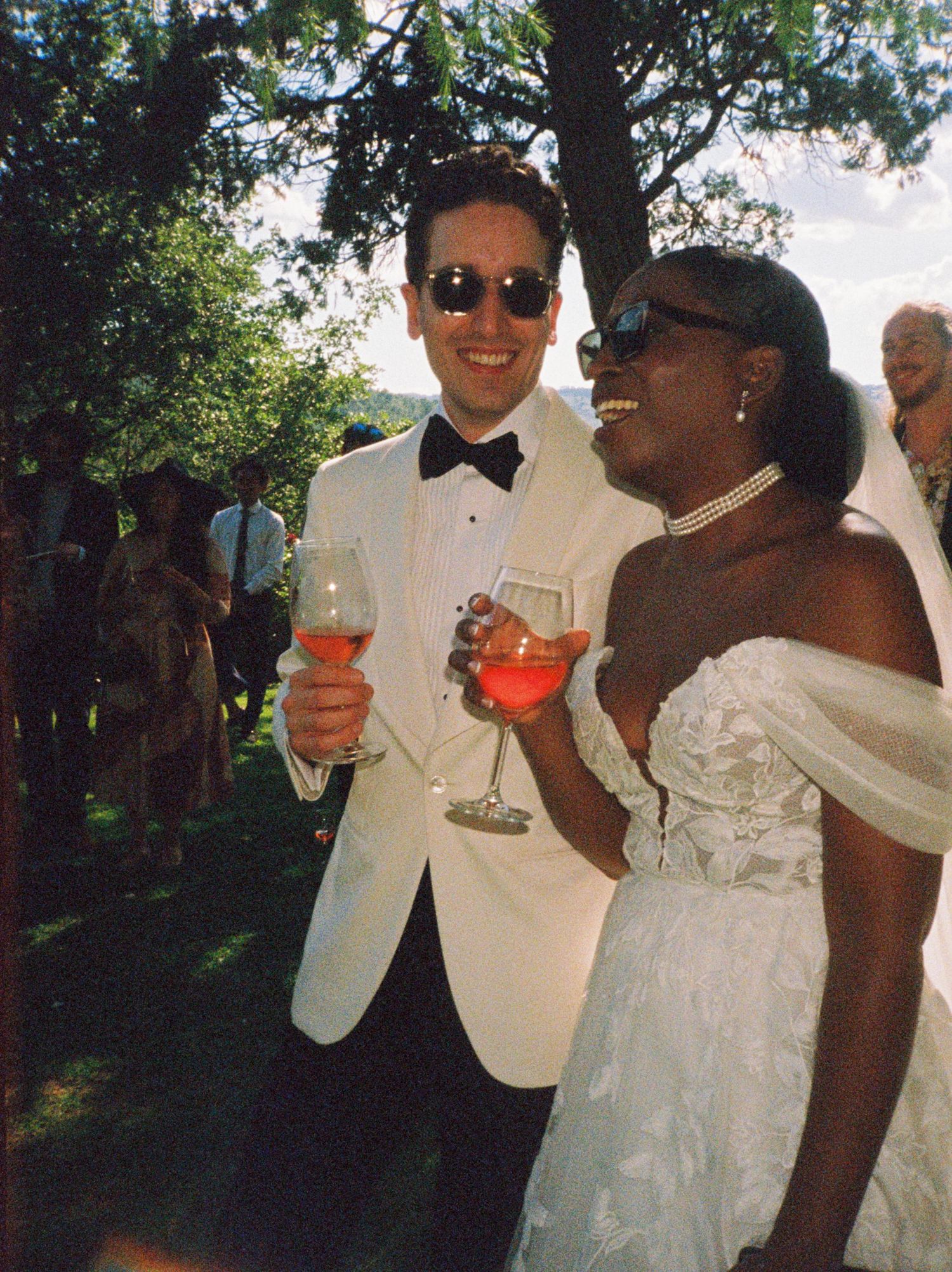 Wedding couple toasts with vibrant cocktails while sharing joyful moments outdoors in formal white attire.