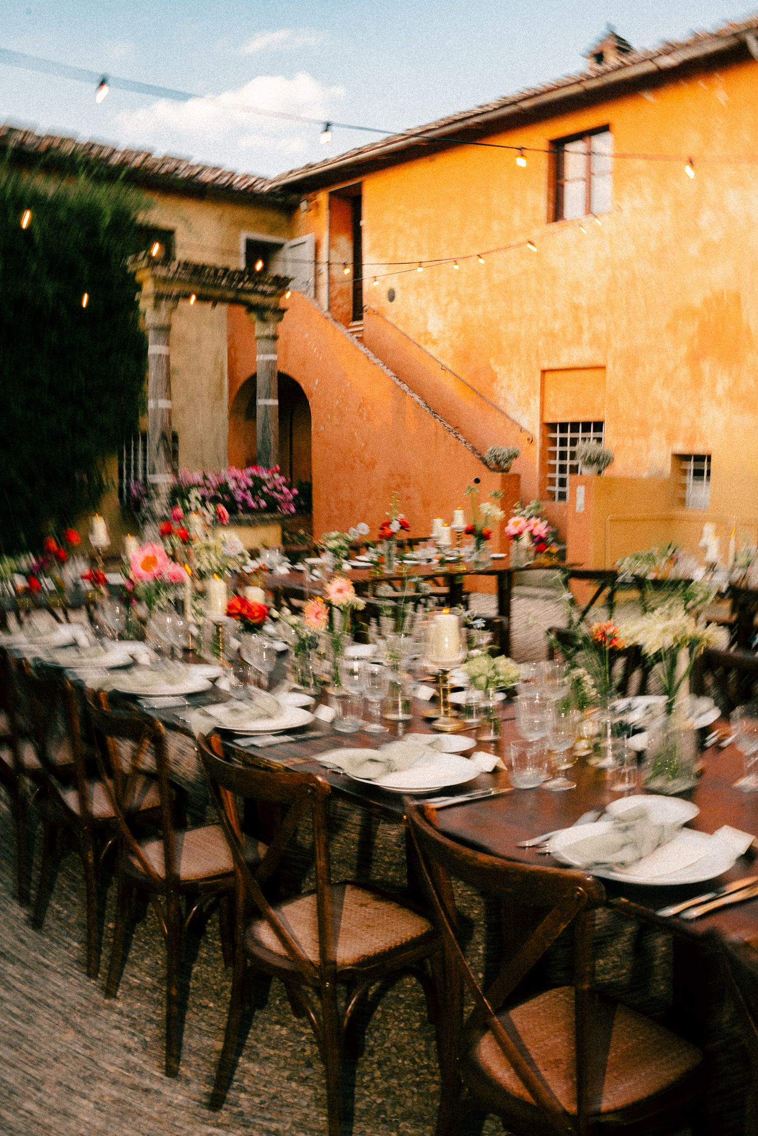 Elegant outdoor wedding reception with long wooden tables, colorful floral arrangements, string lights, and rustic Tuscan villa backdrop.