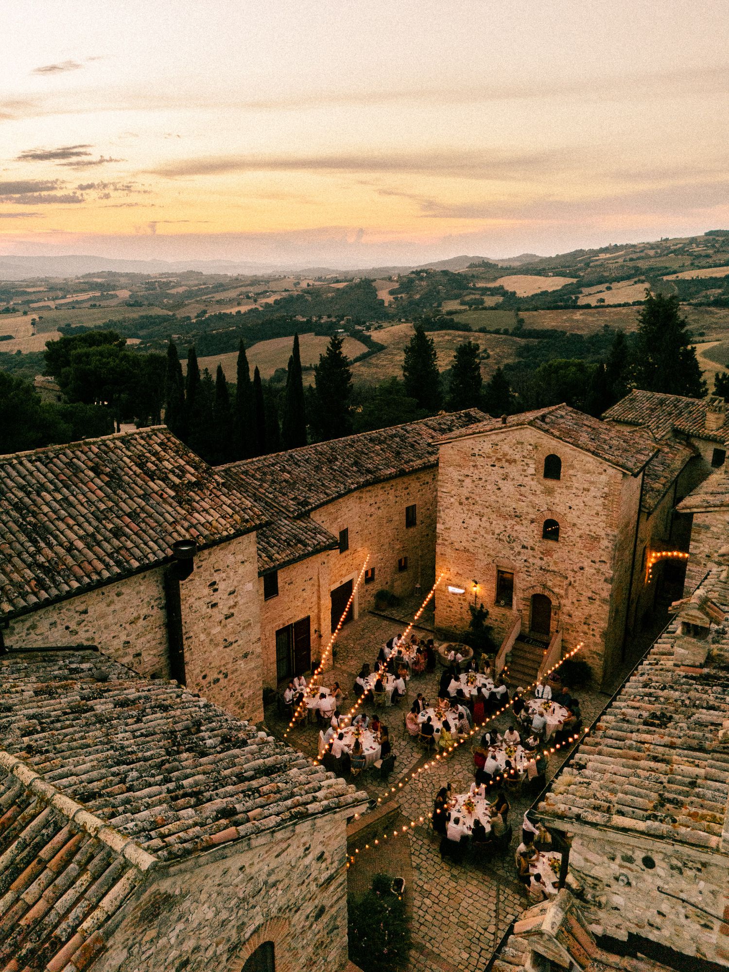 Aerial view of a Tuscan stone villa courtyard wedding reception at sunset with string lights and rolling hills backdrop.