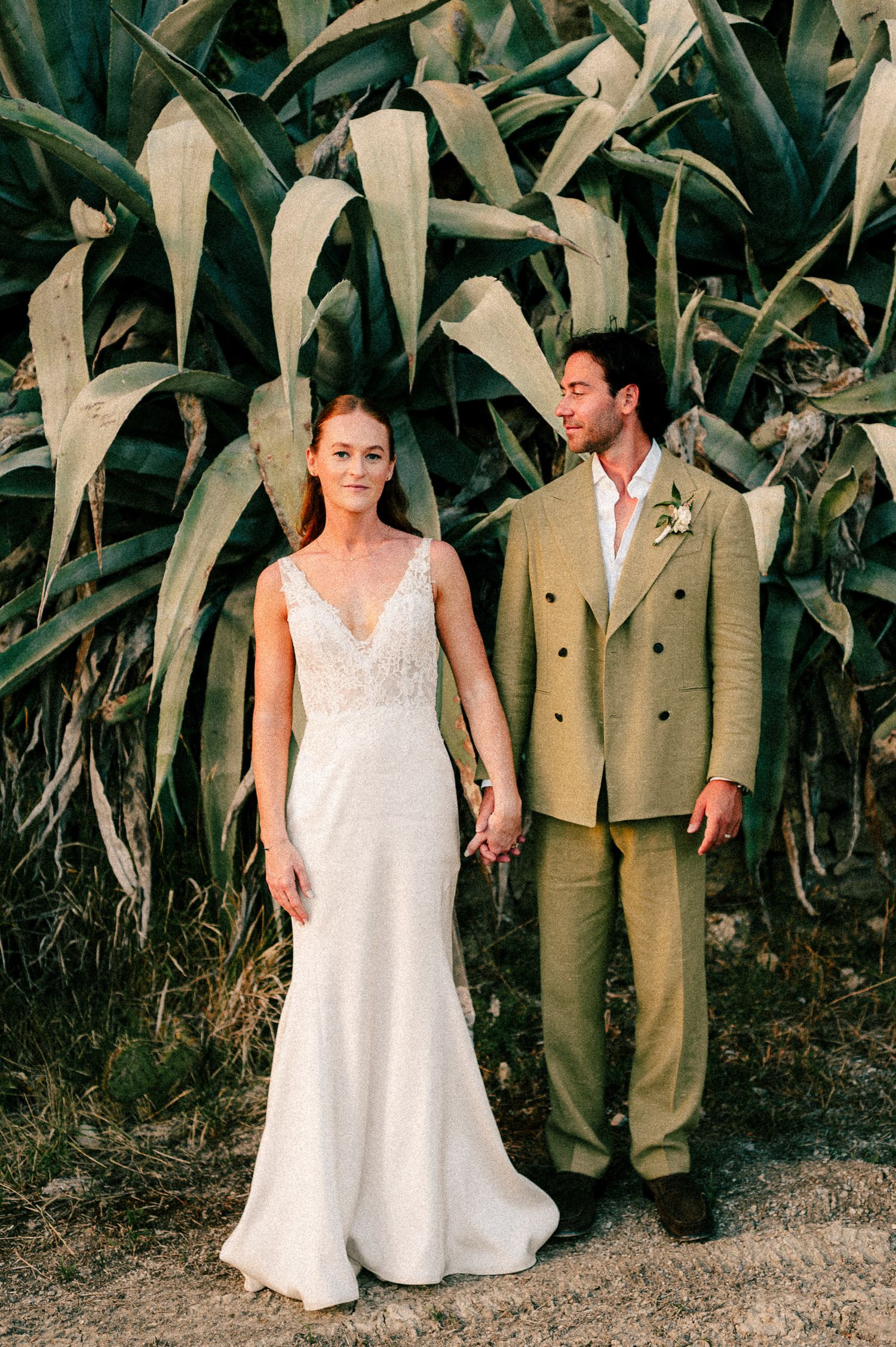 Bride in white lace gown and groom in olive suit hold hands before lush agave plants at outdoor wedding.