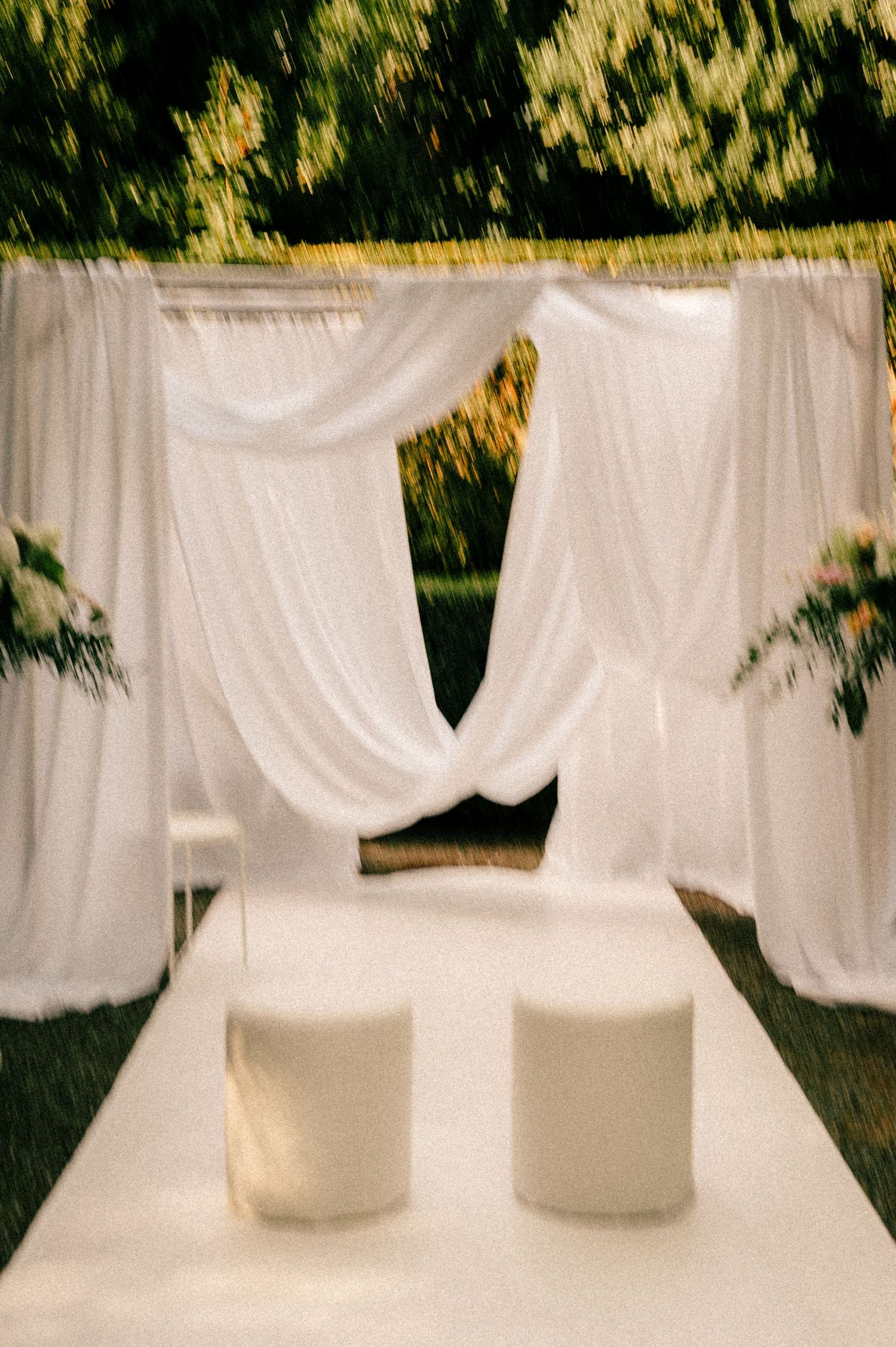 Elegant outdoor wedding chuppah with white draped fabric, floral arrangements, and two white ottomans on an aisle runner.