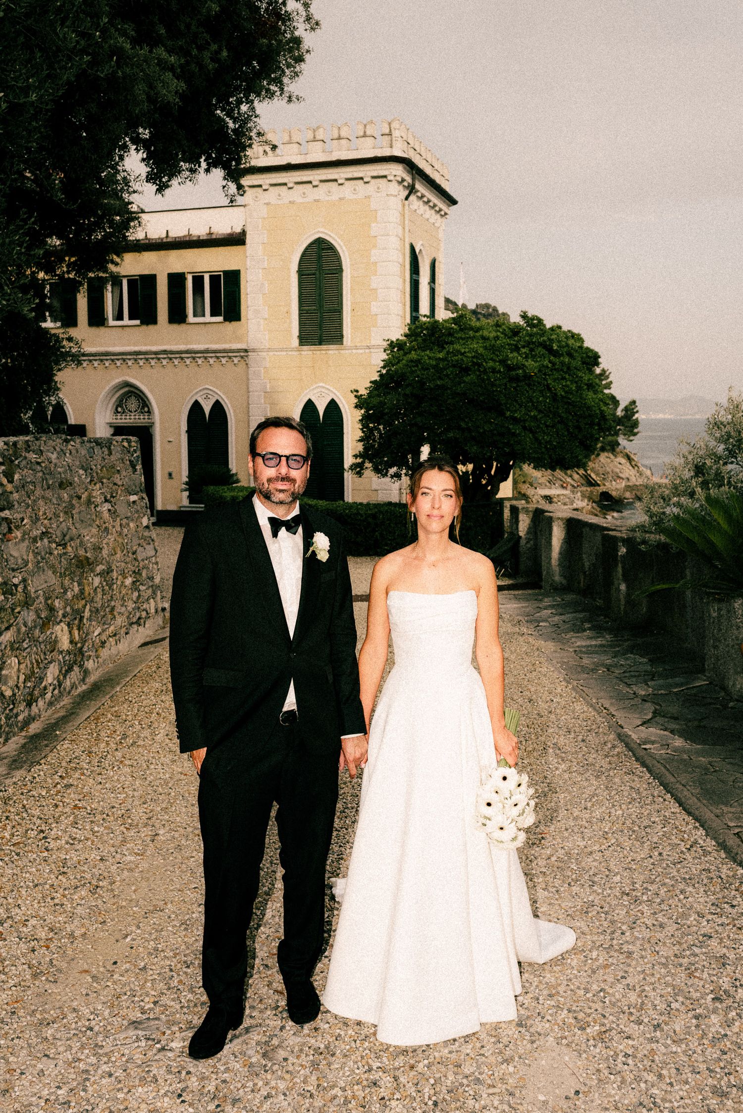 Bride and groom holding hands in front of a historic villa on their wedding day, bride holding white bouquet.