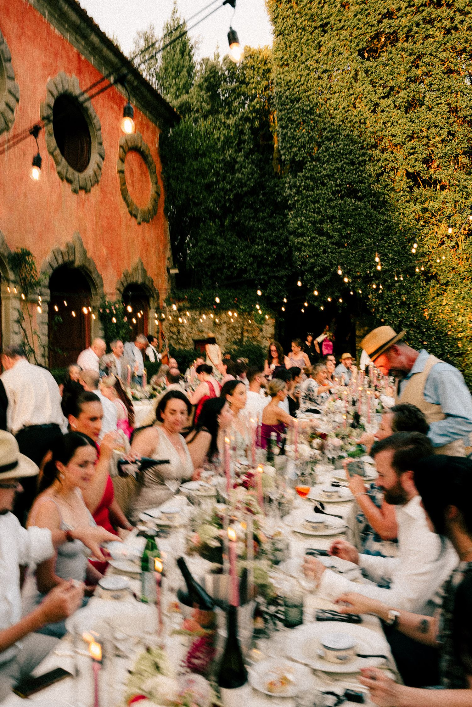 Guests dining at a long outdoor table adorned with string lights, surrounded by lush greenery and a rustic red building.