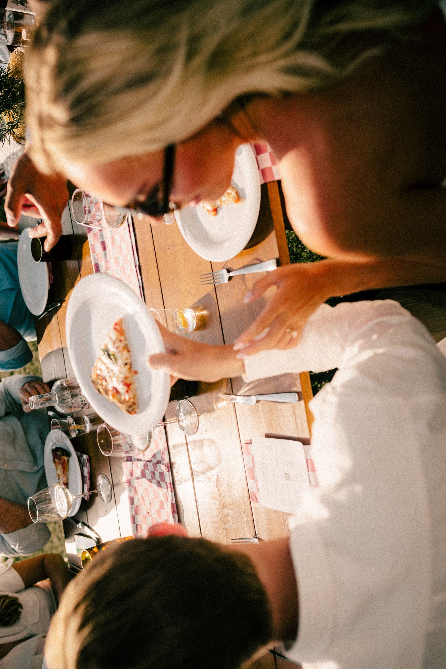 Aerial view of a blonde woman serving food onto white plates at an outdoor dining table with glasses.
