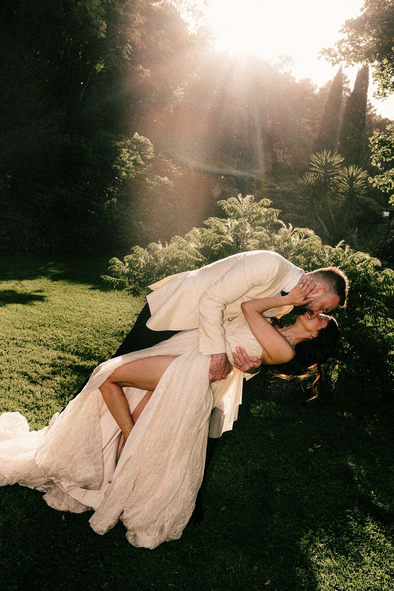 Bride and groom share a romantic dip kiss in a sunlit garden, surrounded by lush greenery on their wedding day.