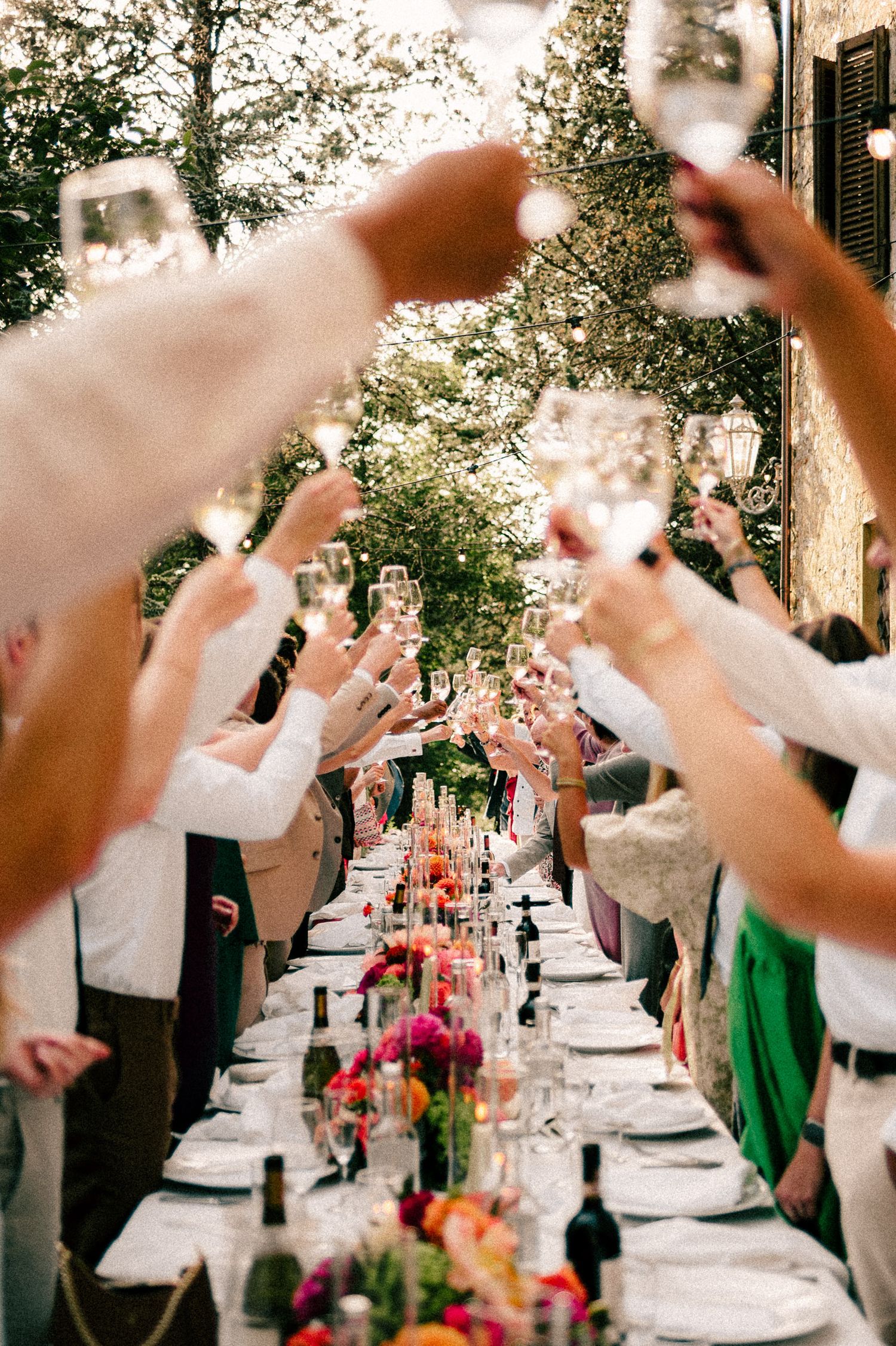 Guests raise champagne glasses in a toast over a long floral decorated outdoor dinner table at an elegant wedding reception.