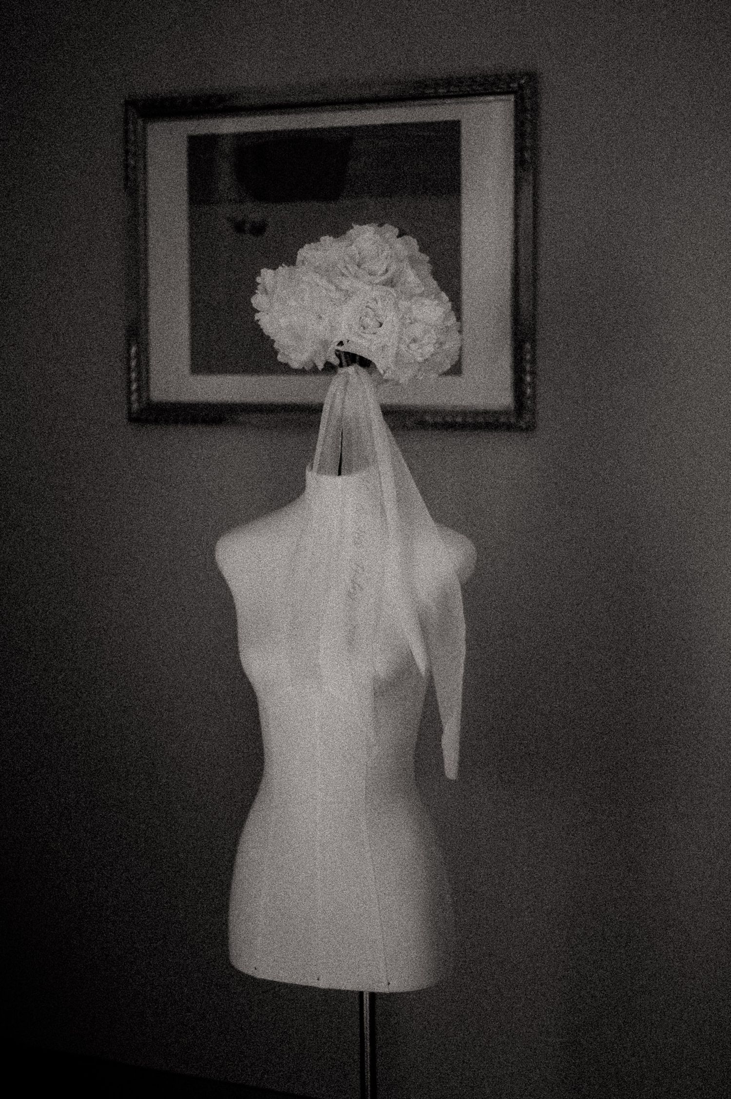 Black and white photo of a dress form mannequin displaying a bridal veil and bouquet of white roses near a framed picture.