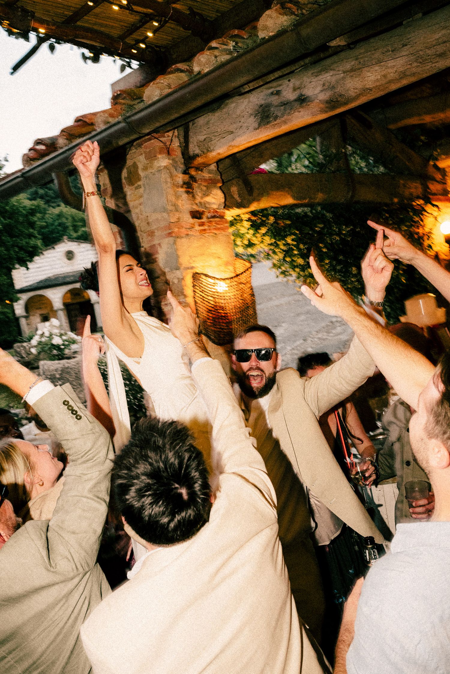 Bride and groom celebrate joyfully with guests at an outdoor rustic wedding reception under wooden beams.
