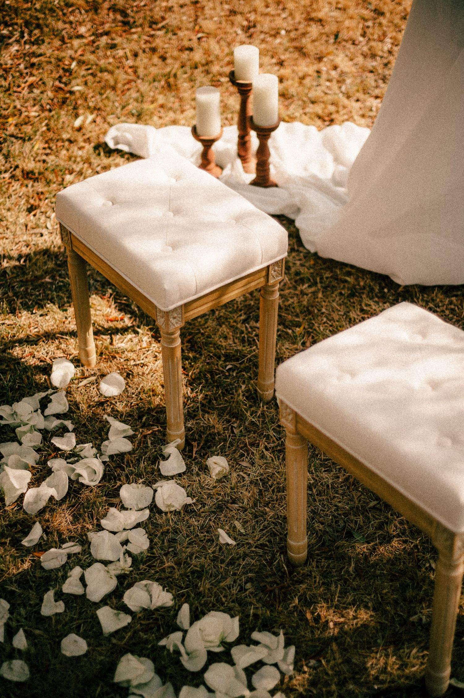 Two white tufted gold-legged stools on grass with rose petals, candles on wooden holder, elegant wedding ceremony decor.