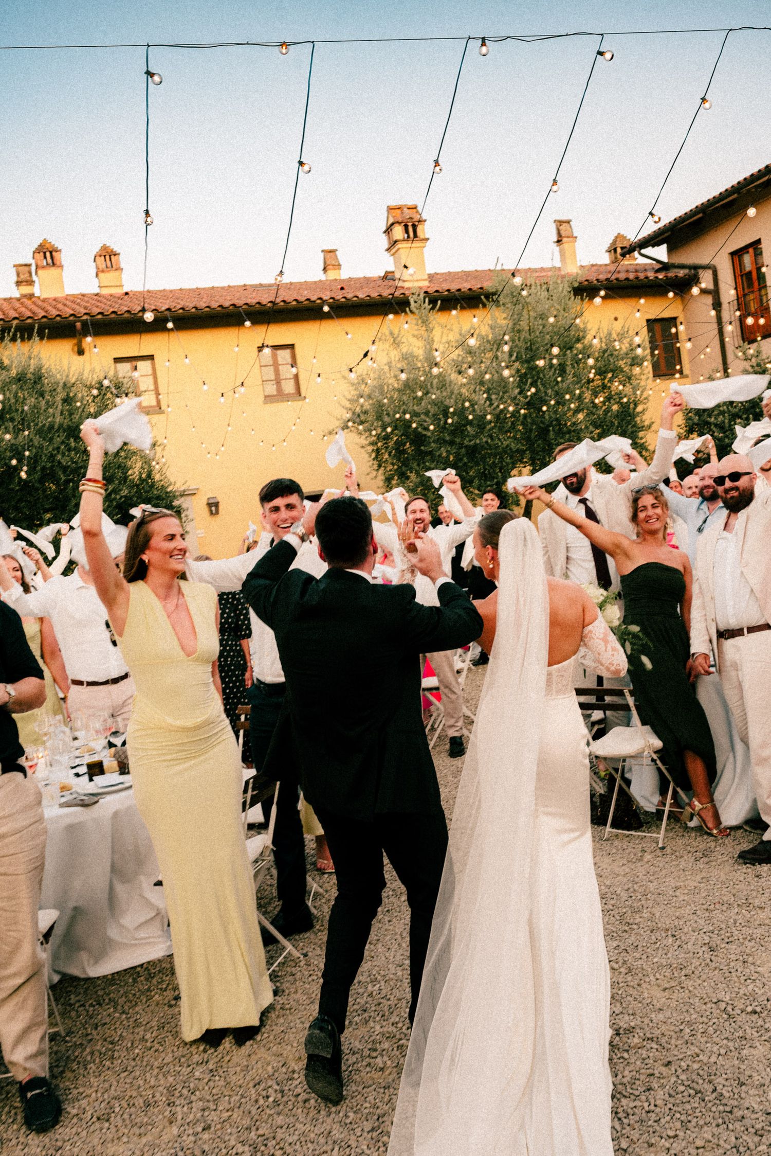 Bride and groom celebrate with guests at an outdoor Italian villa wedding reception with string lights at dusk.