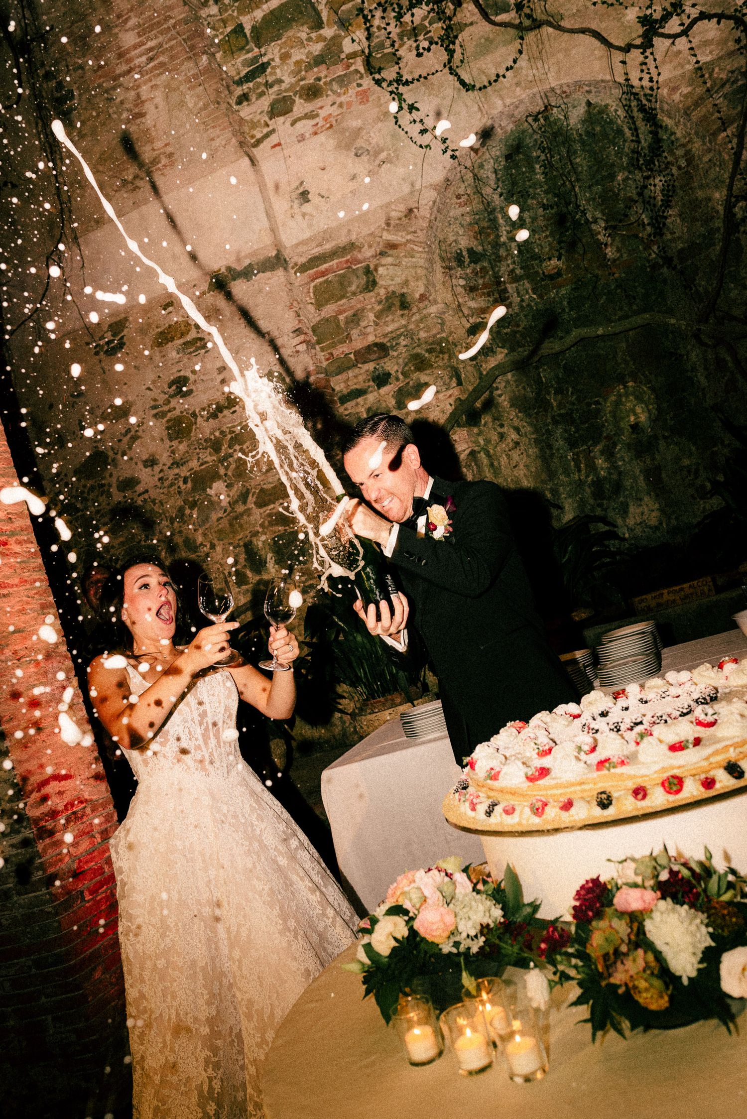 Bride and groom celebrating with champagne spray beside wedding cake in rustic brick venue.