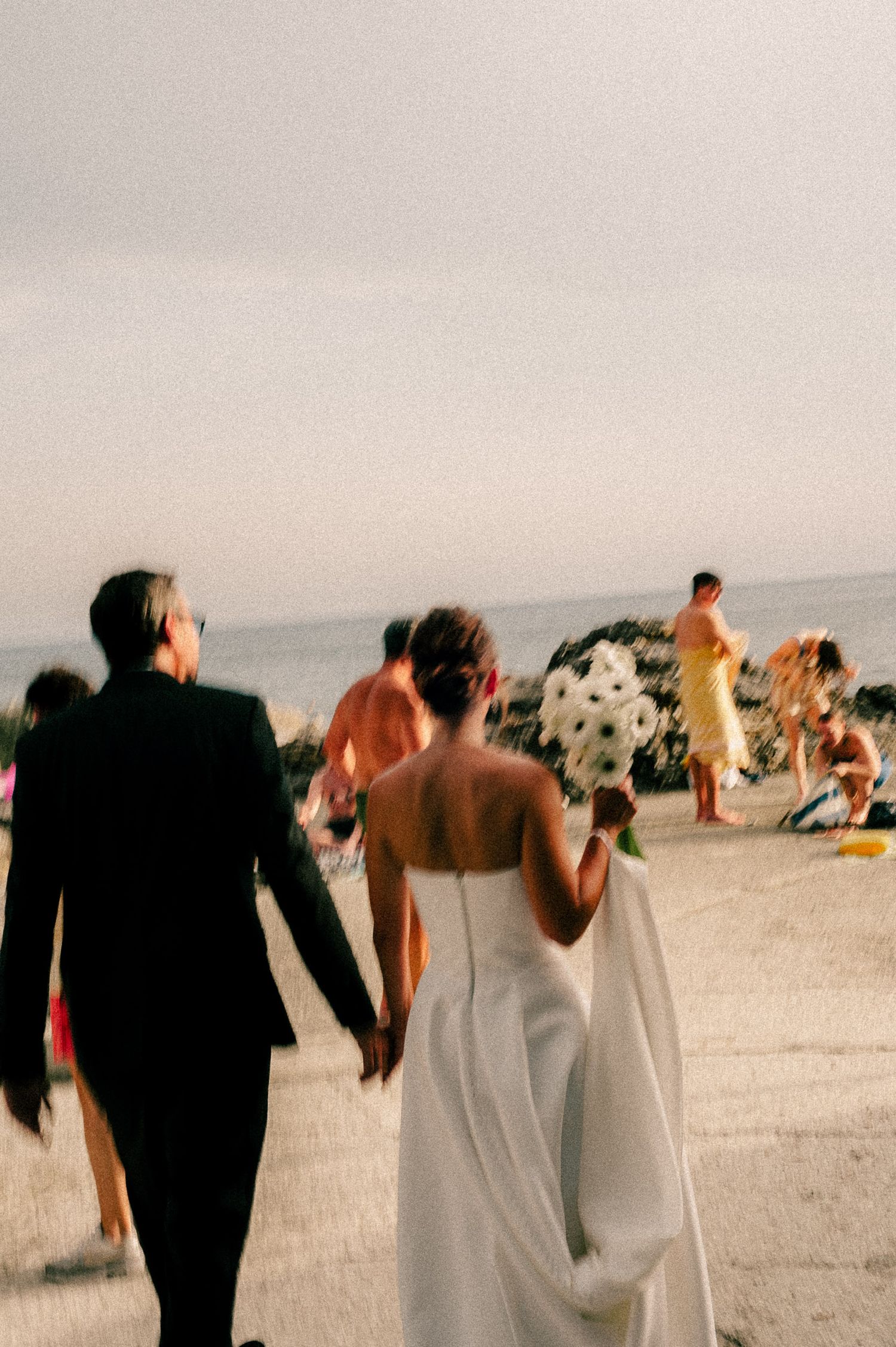 A bride and groom walk hand-in-hand on a beach, she holds a black and white bouquet, guests gather near the shore.