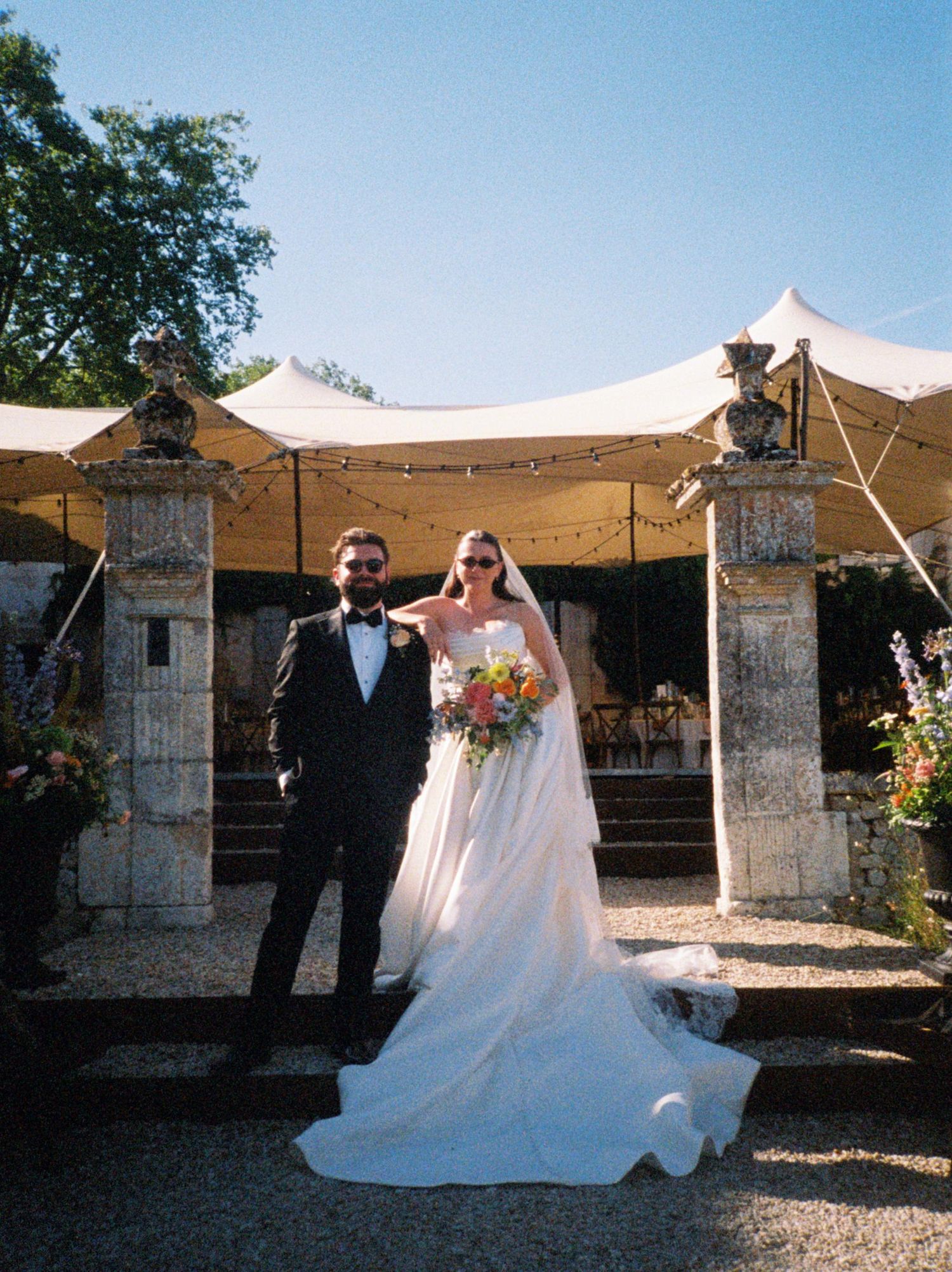 Bride and groom posing between stone pillars at an outdoor wedding venue with a white tent and string lights.