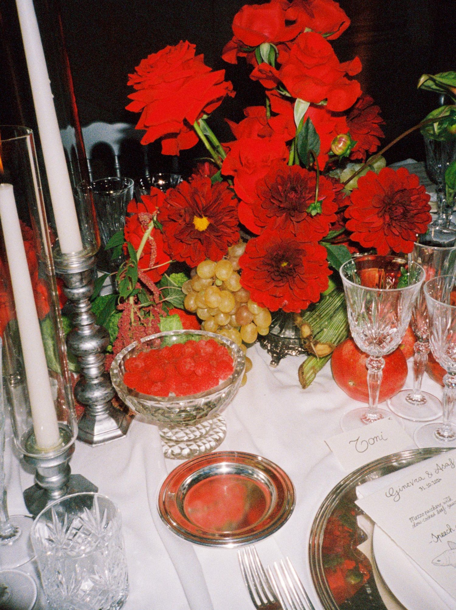 Elegant red floral centerpiece with dahlias, candles, crystal glasses, and silver dishes on a white tablecloth.