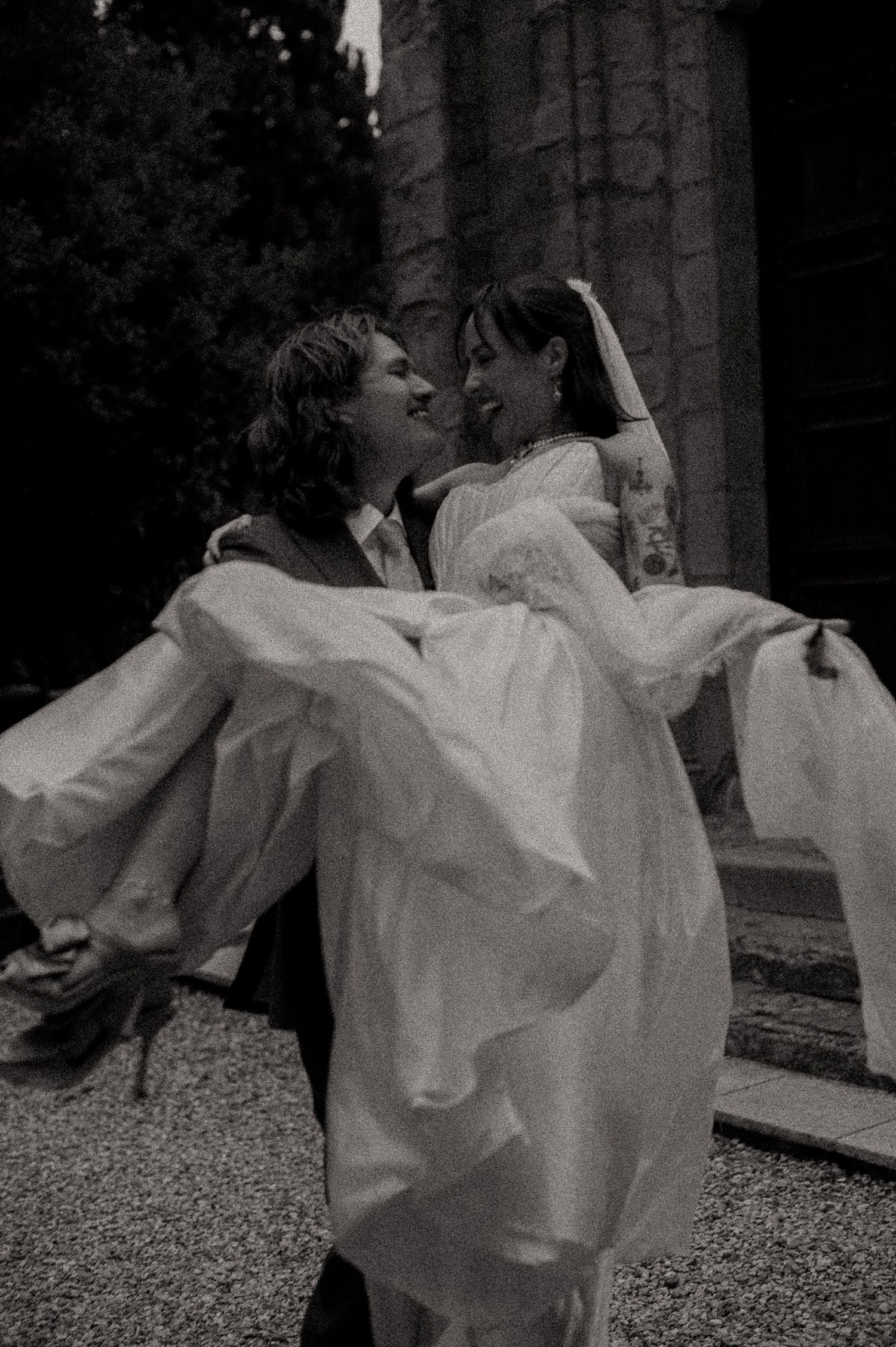 Black and white photo of a groom carrying his bride in a wedding dress, both smiling joyfully outside a church.