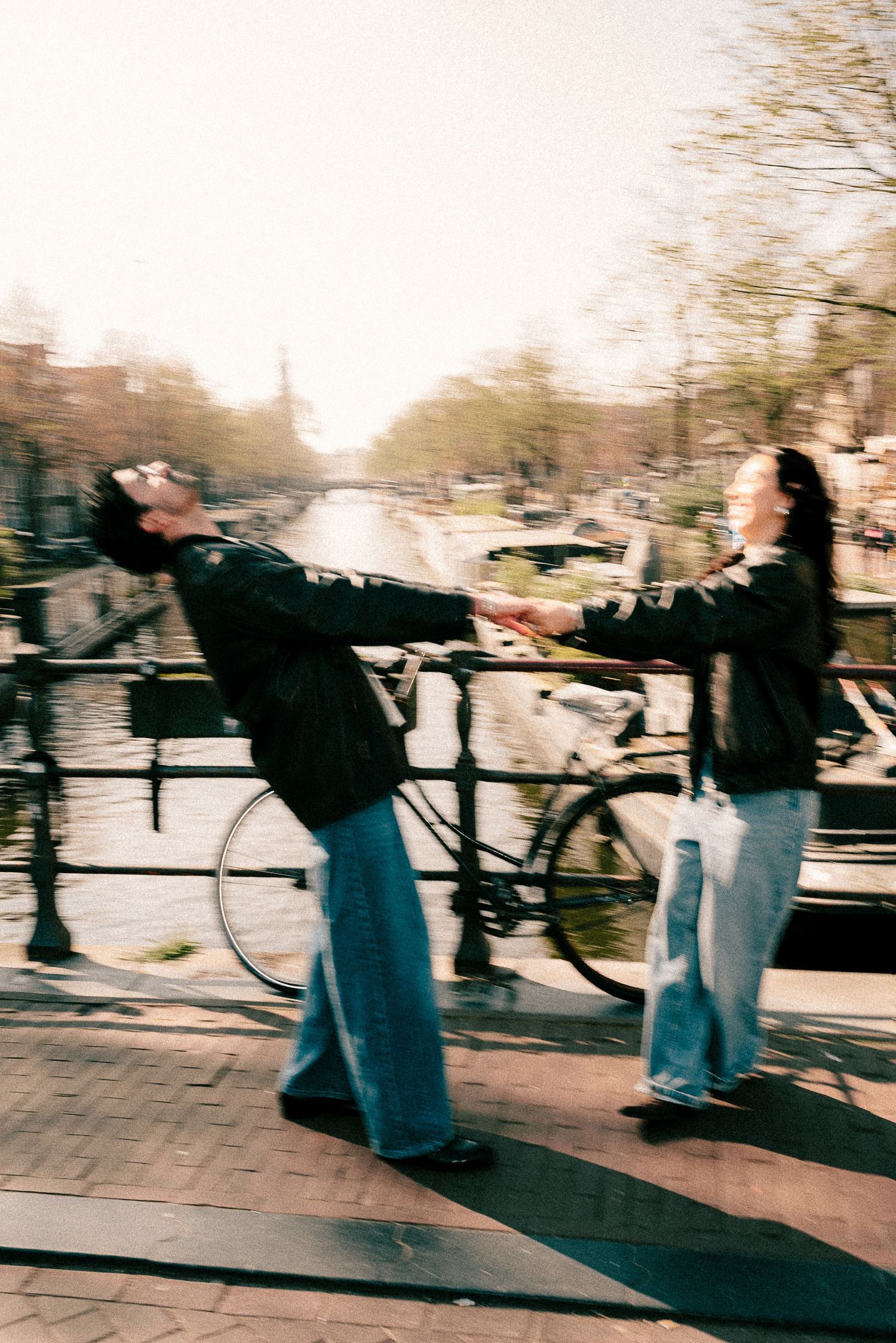 Two people holding hands and dancing on a bridge over an Amsterdam canal, with bicycles and trees in the background.