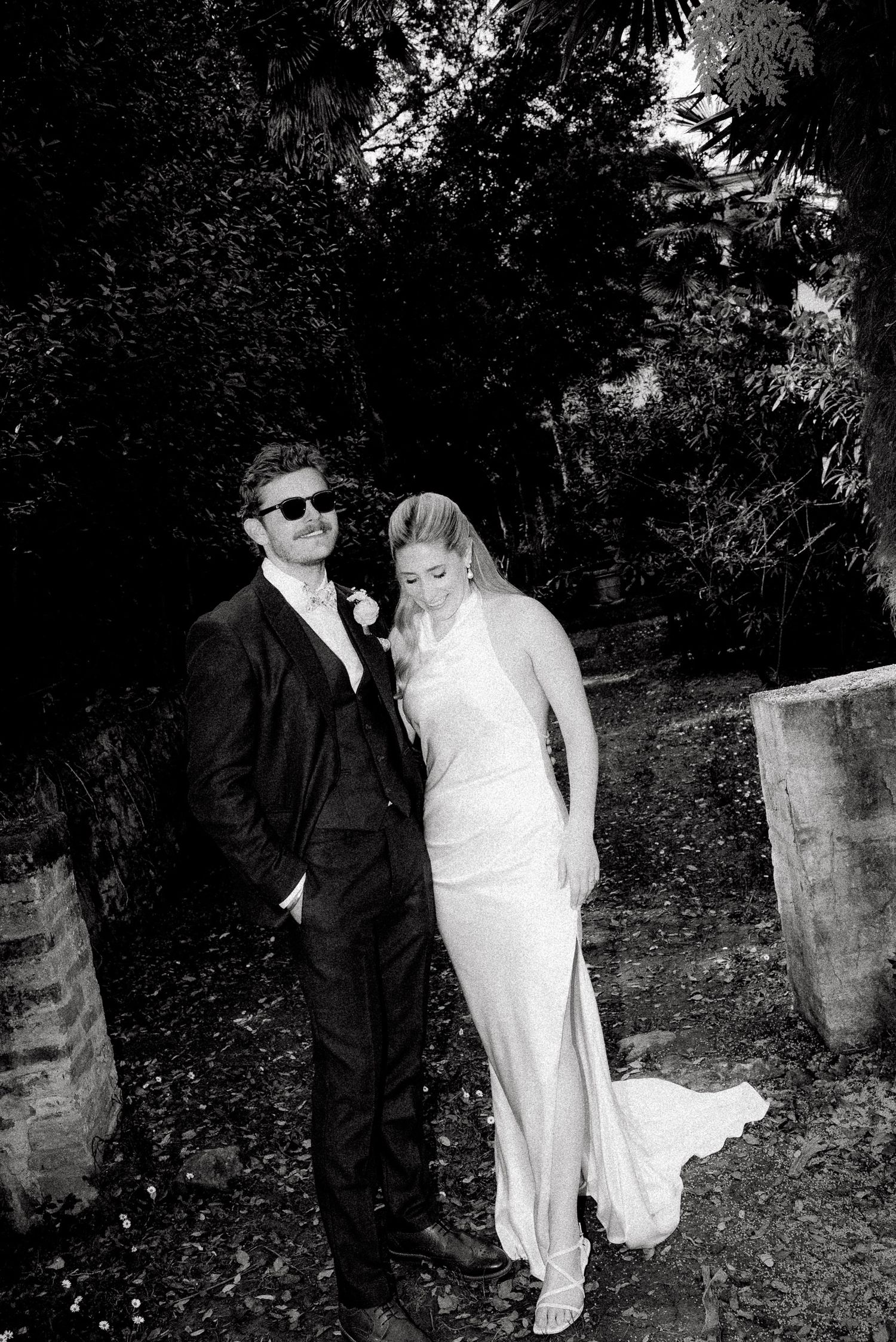 Black and white wedding photo of bride in white gown and groom in dark suit with sunglasses posing outdoors.