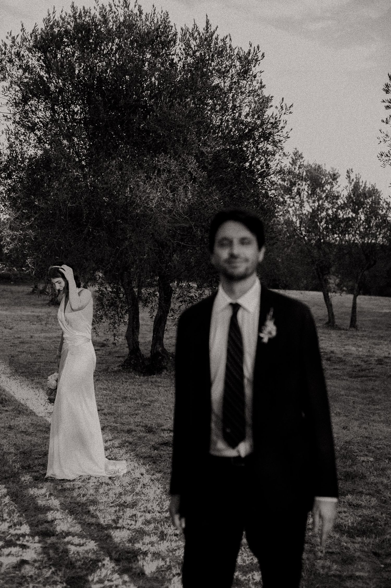 Black and white wedding photo of groom in suit with boutonniere standing foreground, bride in white gown posed near trees background.