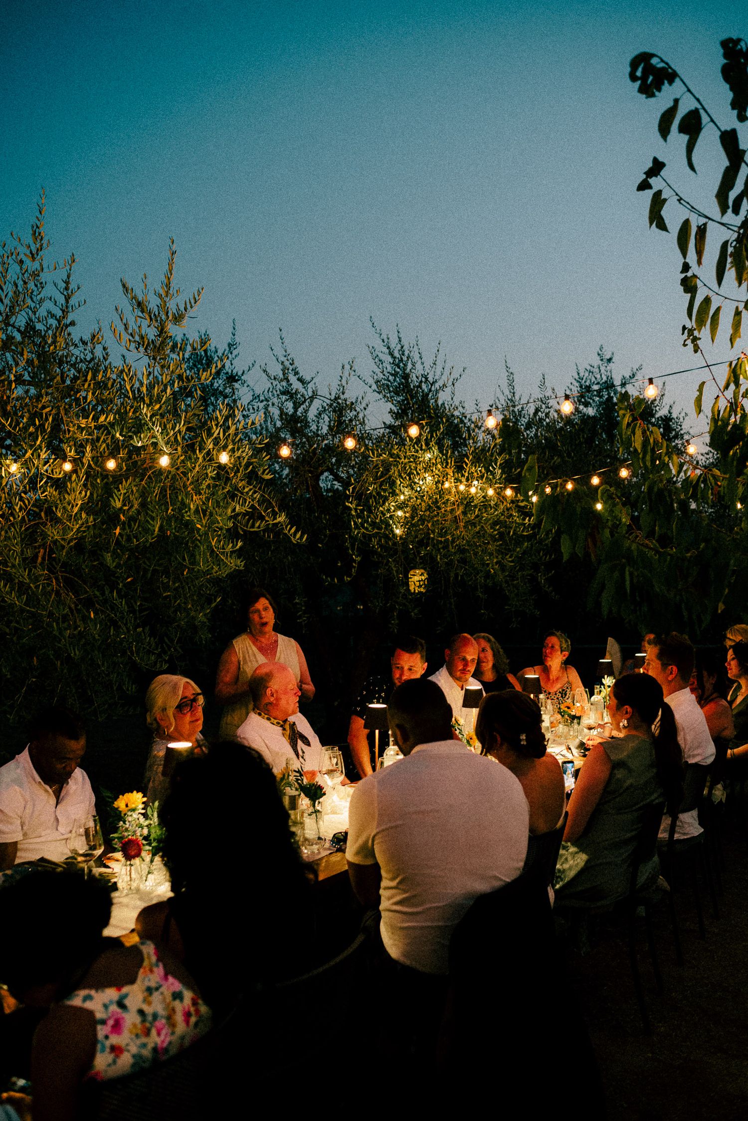 Guests dining outdoors at dusk under string lights surrounded by lush greenery at an elegant evening garden party.