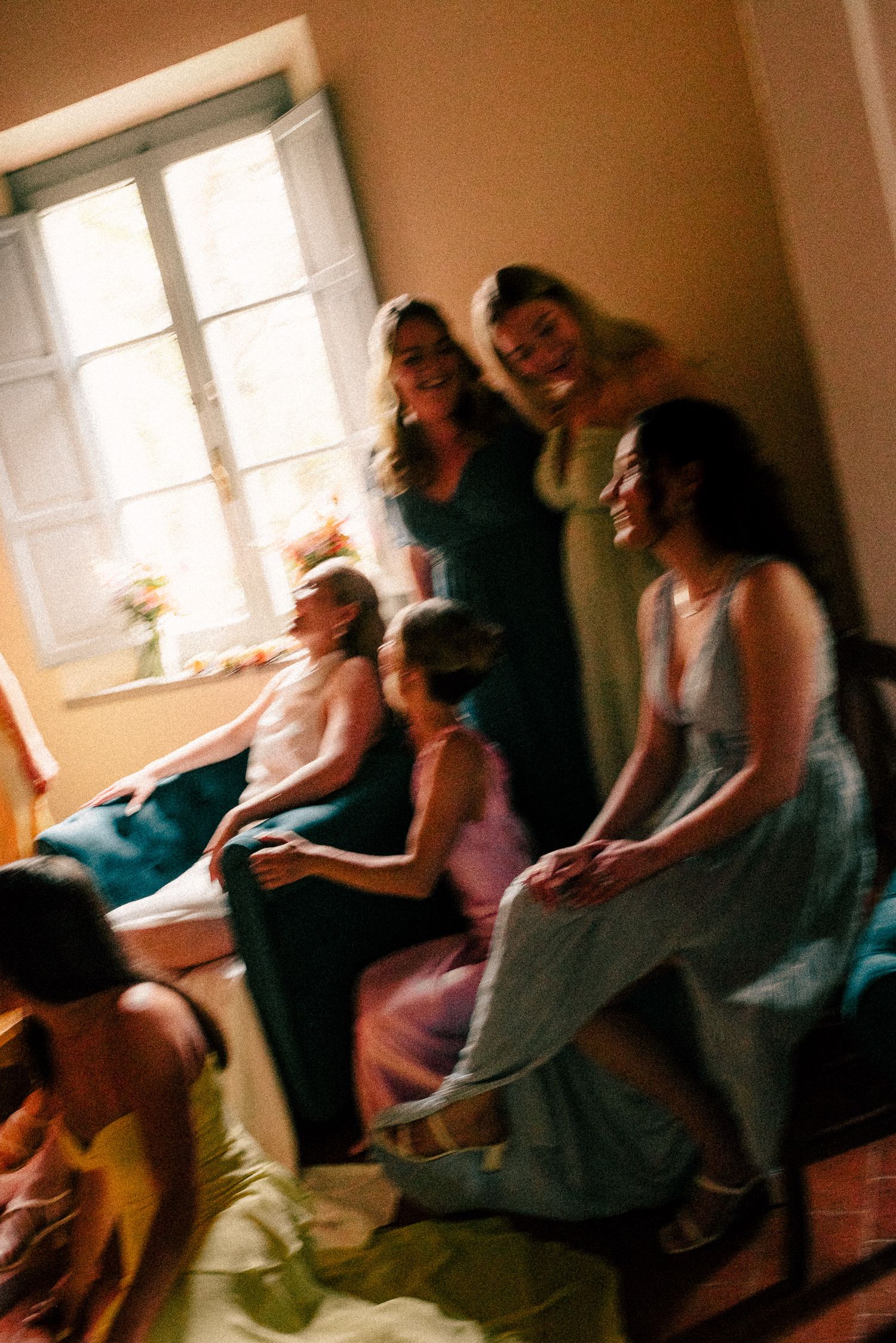 Group of women in colorful formal dresses laughing together in a warmly lit room near a window.