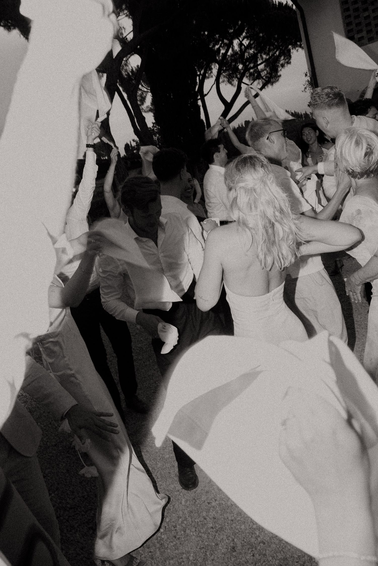 Black and white photo of a bride in white dress dancing joyfully with guests at an outdoor wedding reception.