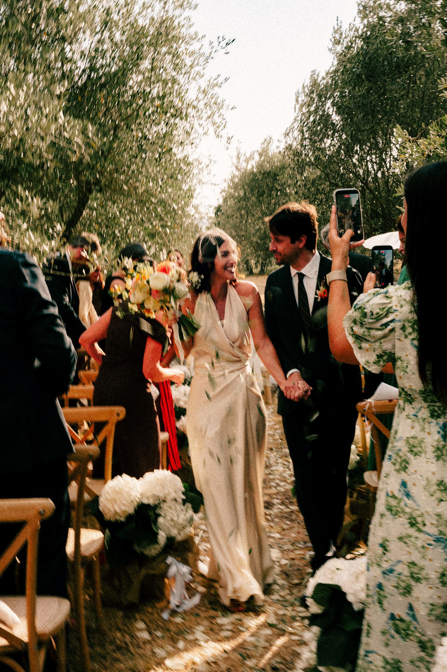 Bride and groom walk down outdoor aisle lined with olive trees, guests cheering and taking photos at rustic wedding ceremony.