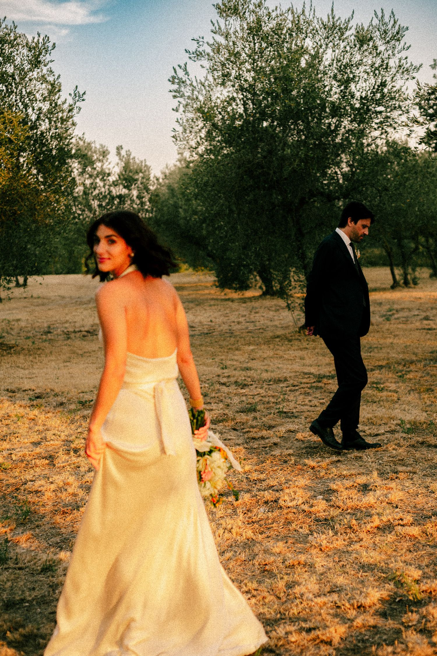 Bride in backless white gown holds bouquet while groom in dark suit walks behind her in an olive grove.