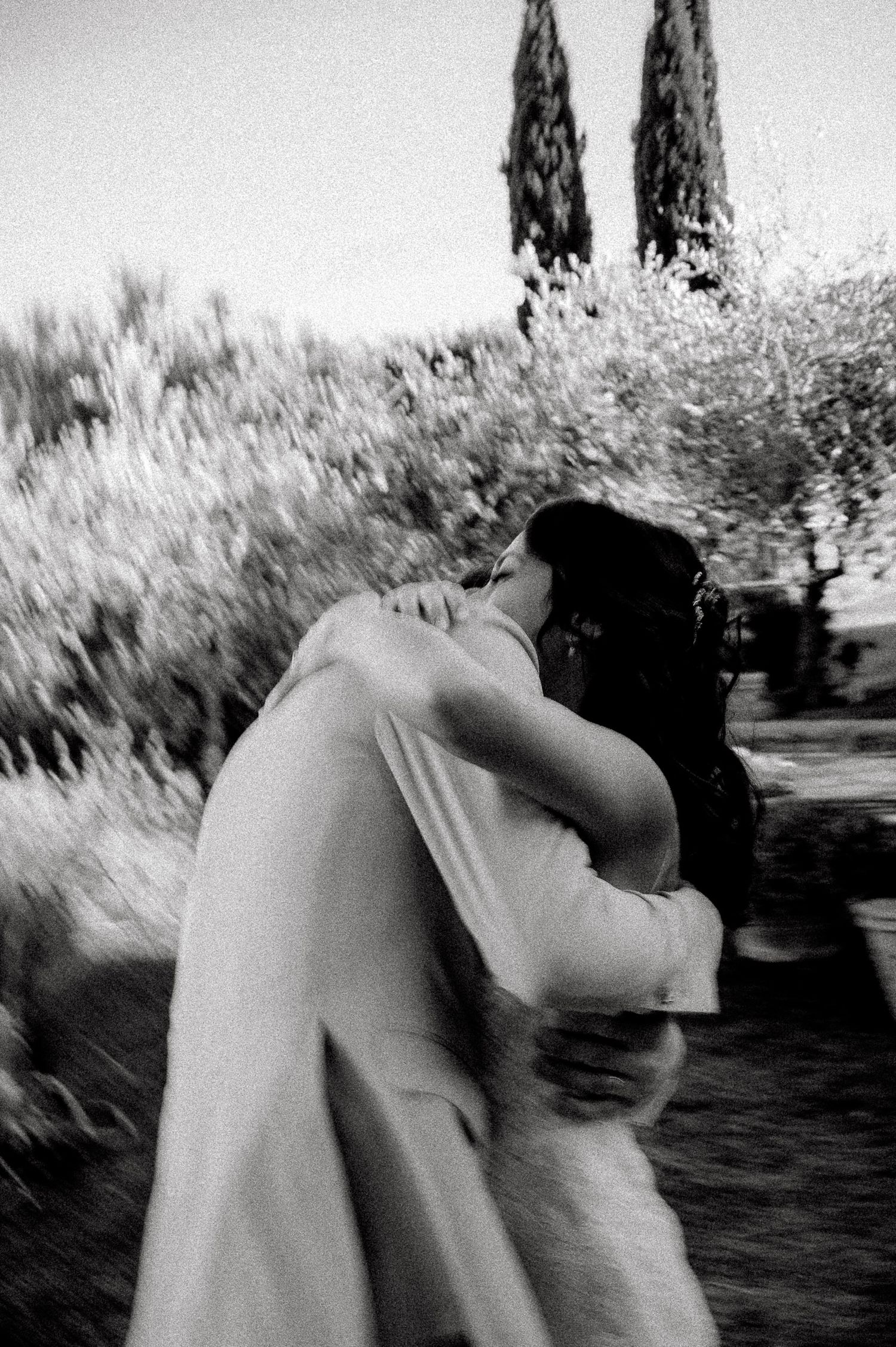 Black and white photo of a couple sharing a romantic kiss outdoors, surrounded by lush trees and cypress in the background.