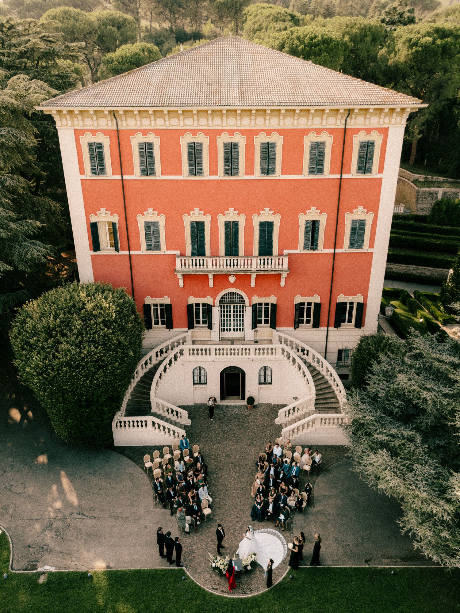Aerial view of a wedding ceremony at a stunning red Italian villa with elegant staircases and lush gardens.