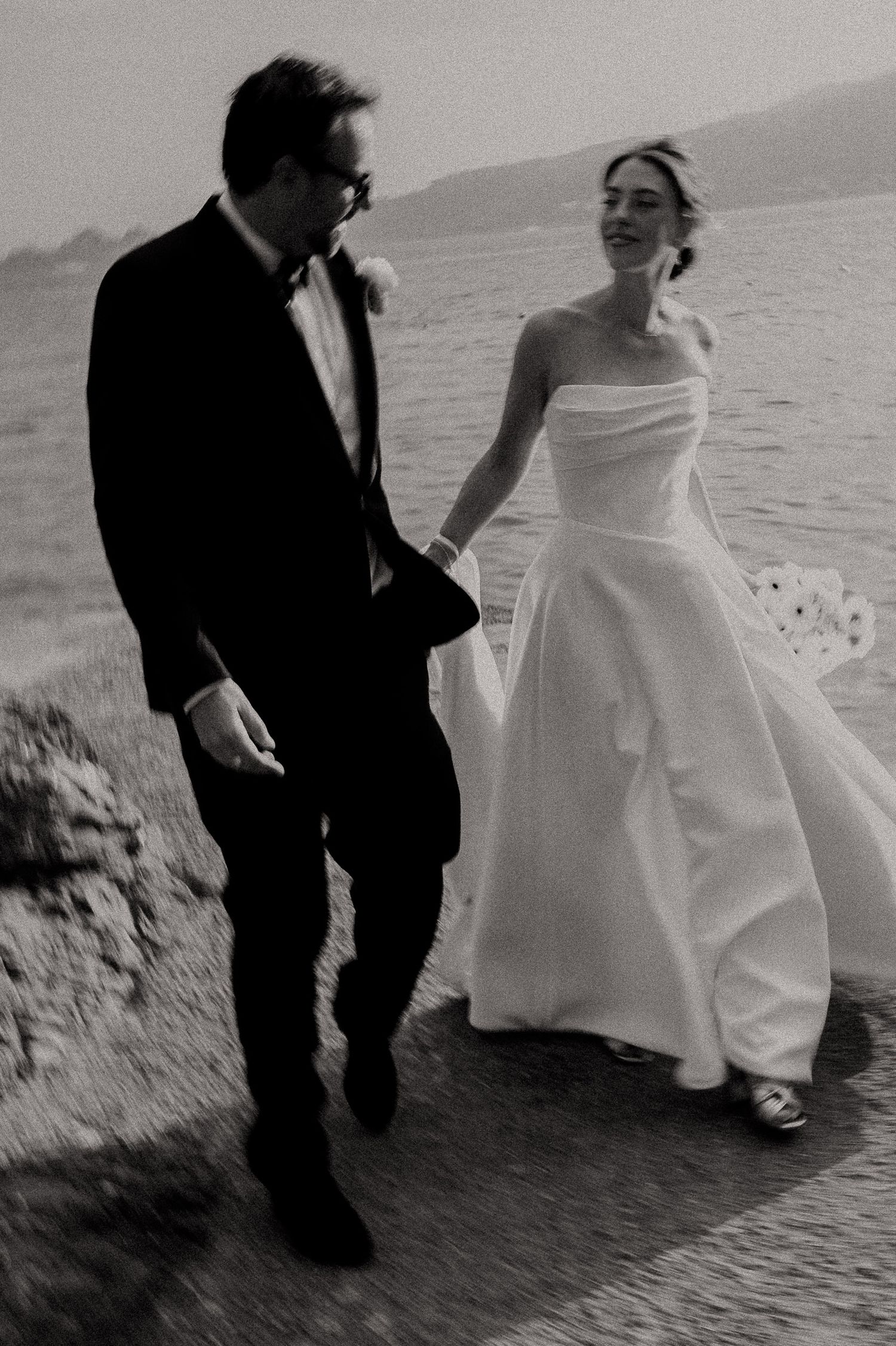 Black and white photo of a bride and groom walking hand in hand by the sea on their wedding day.
