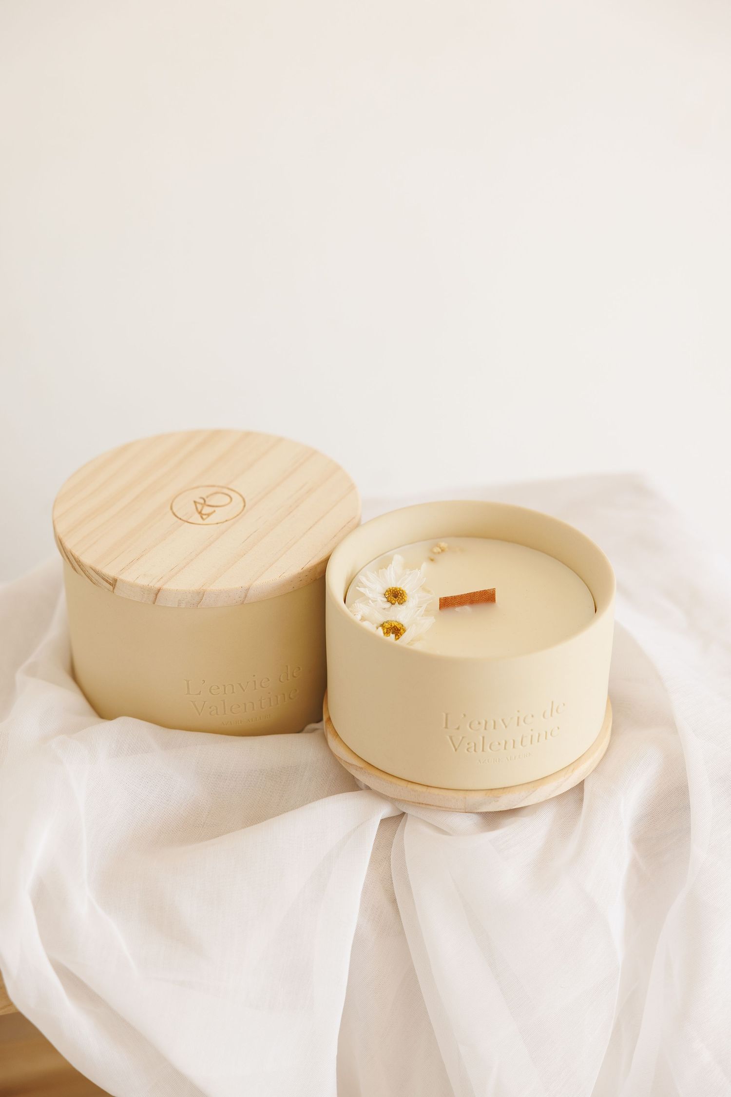 Minimalist cream-colored candle in a ceramic vessel with wooden lid on white fabric background.